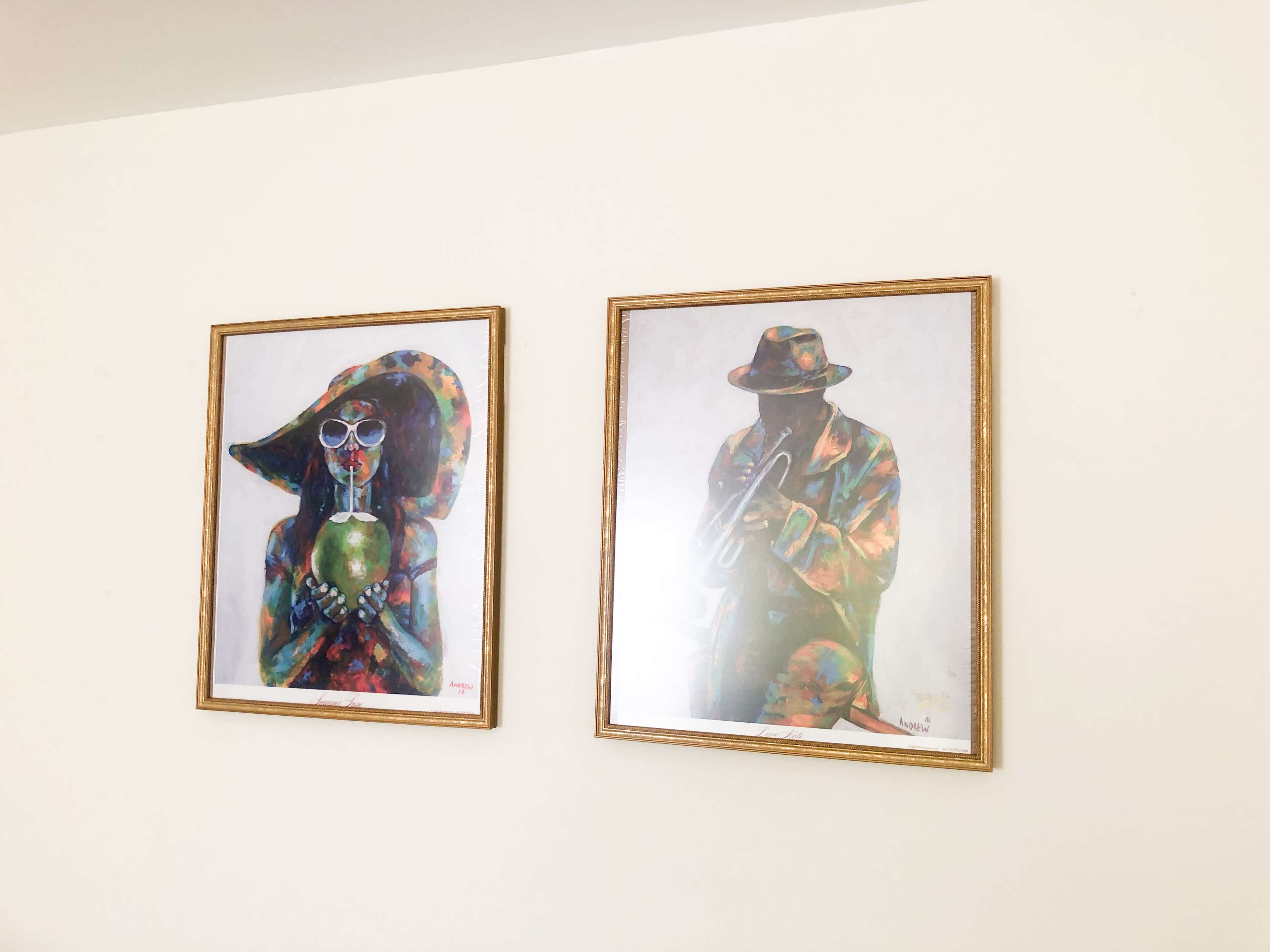 Two framed artworks depicting a woman with a large hat holding a green object and a man playing a trumpet, both featuring vibrant colors, hang on a white wall.