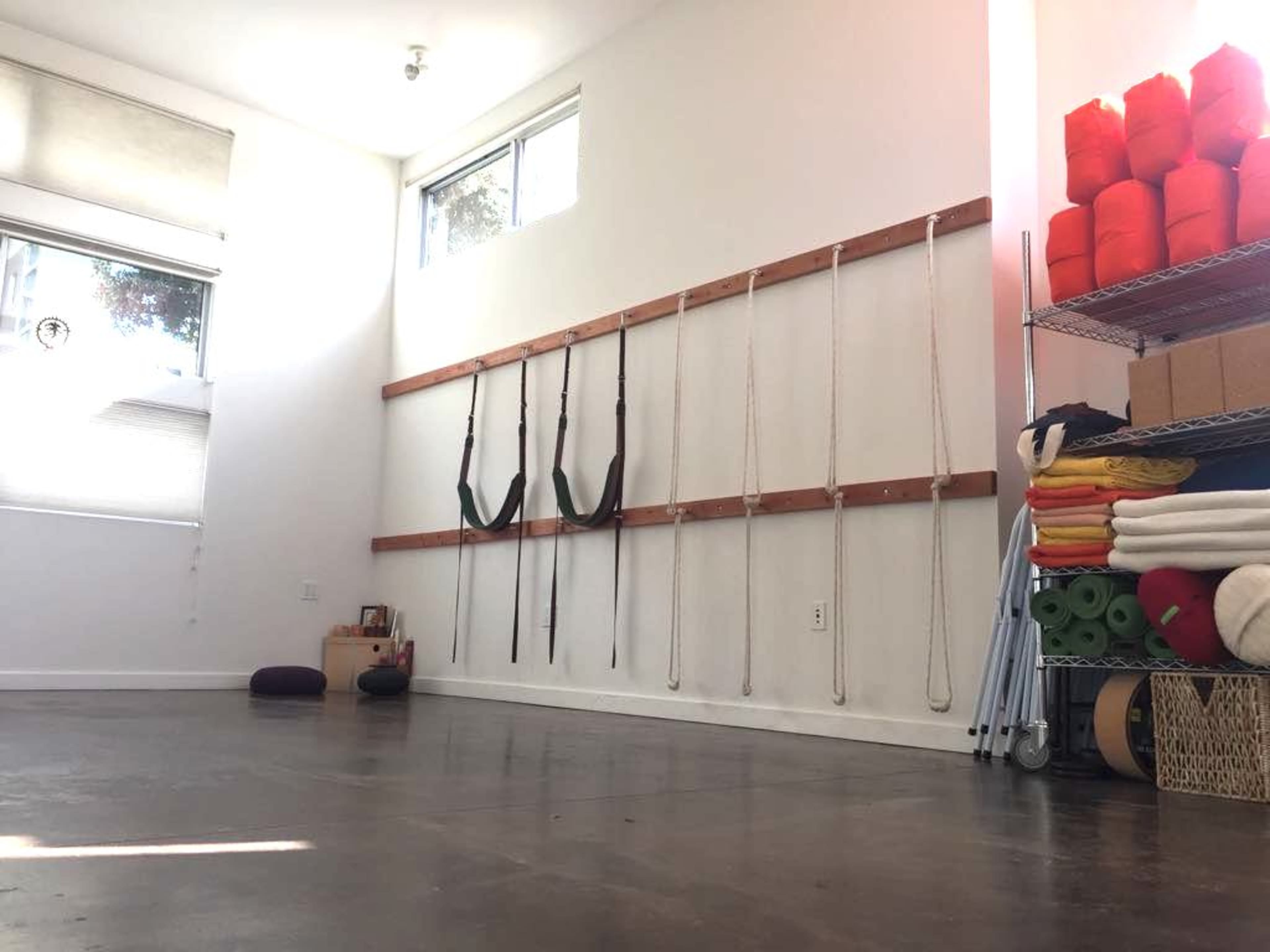 A bright, minimalist room with a wooden wall-mounted system for yoga straps and ropes, alongside neatly organized exercise mats and props stored on shelves.