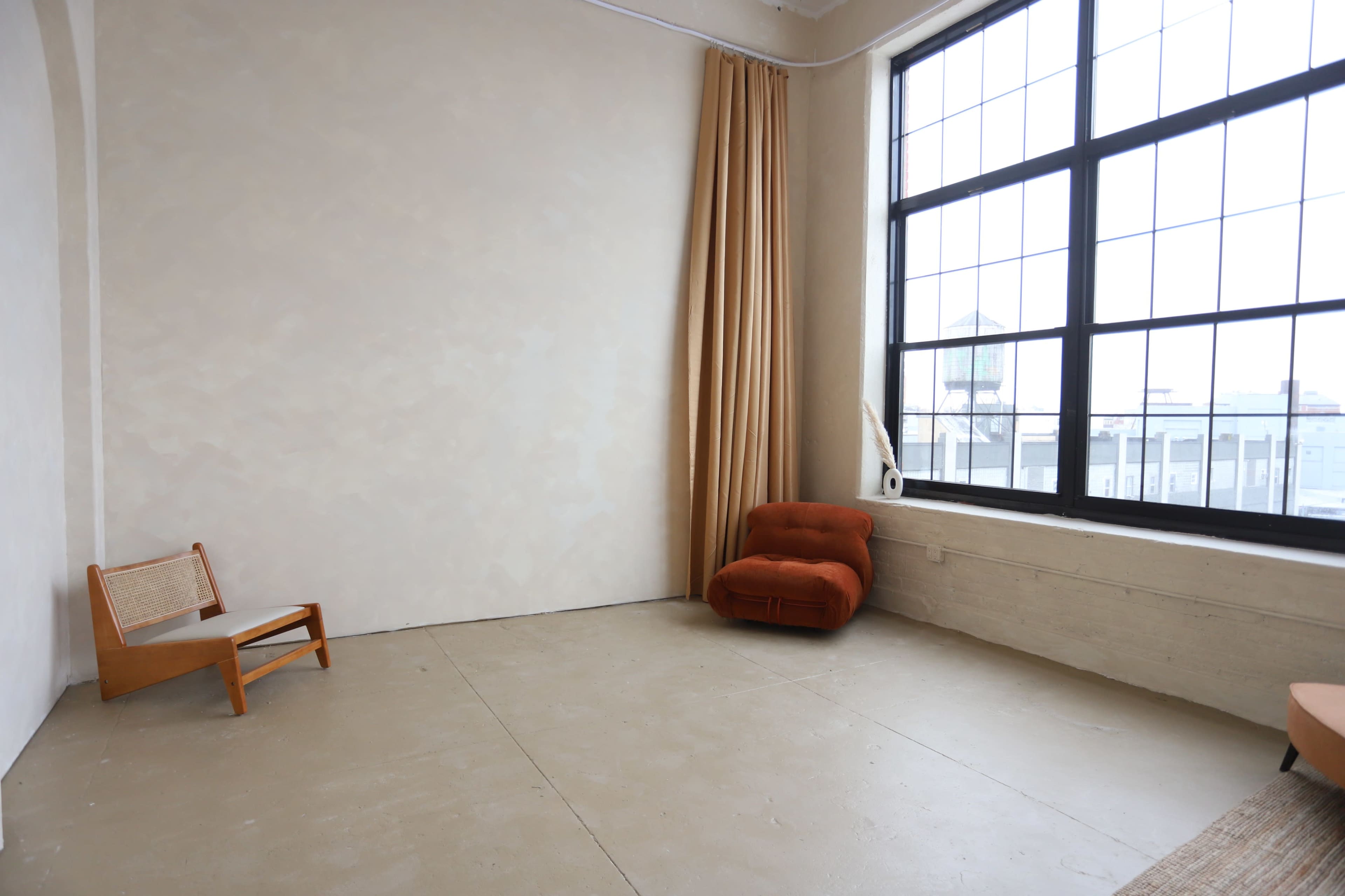 A spacious room with a large window, a wooden chair, and a red upholstered chair against a plain wall.