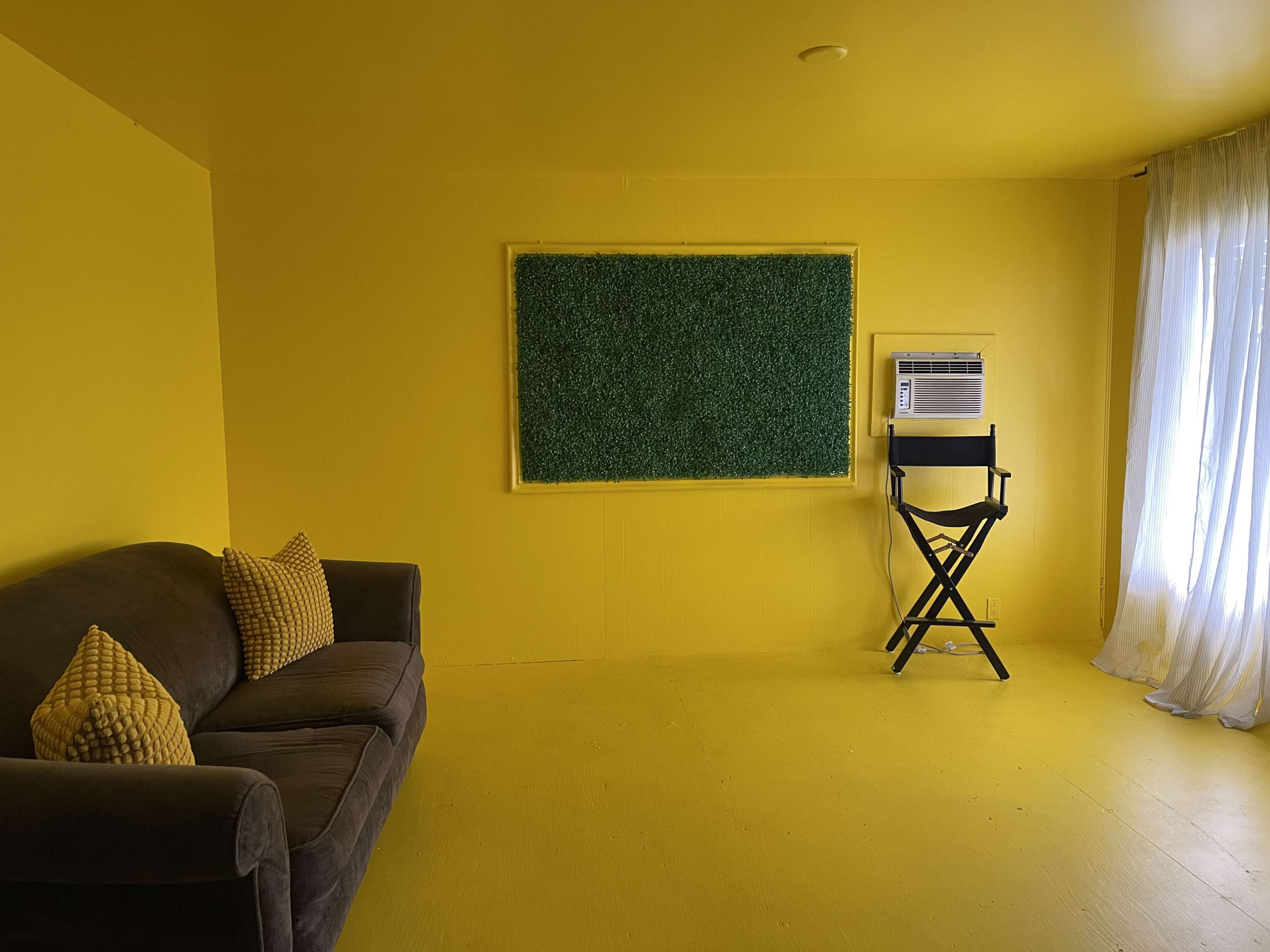 The room features yellow walls and flooring, a gray sofa with yellow pillows, a black director's chair, and a green framed board on one wall.