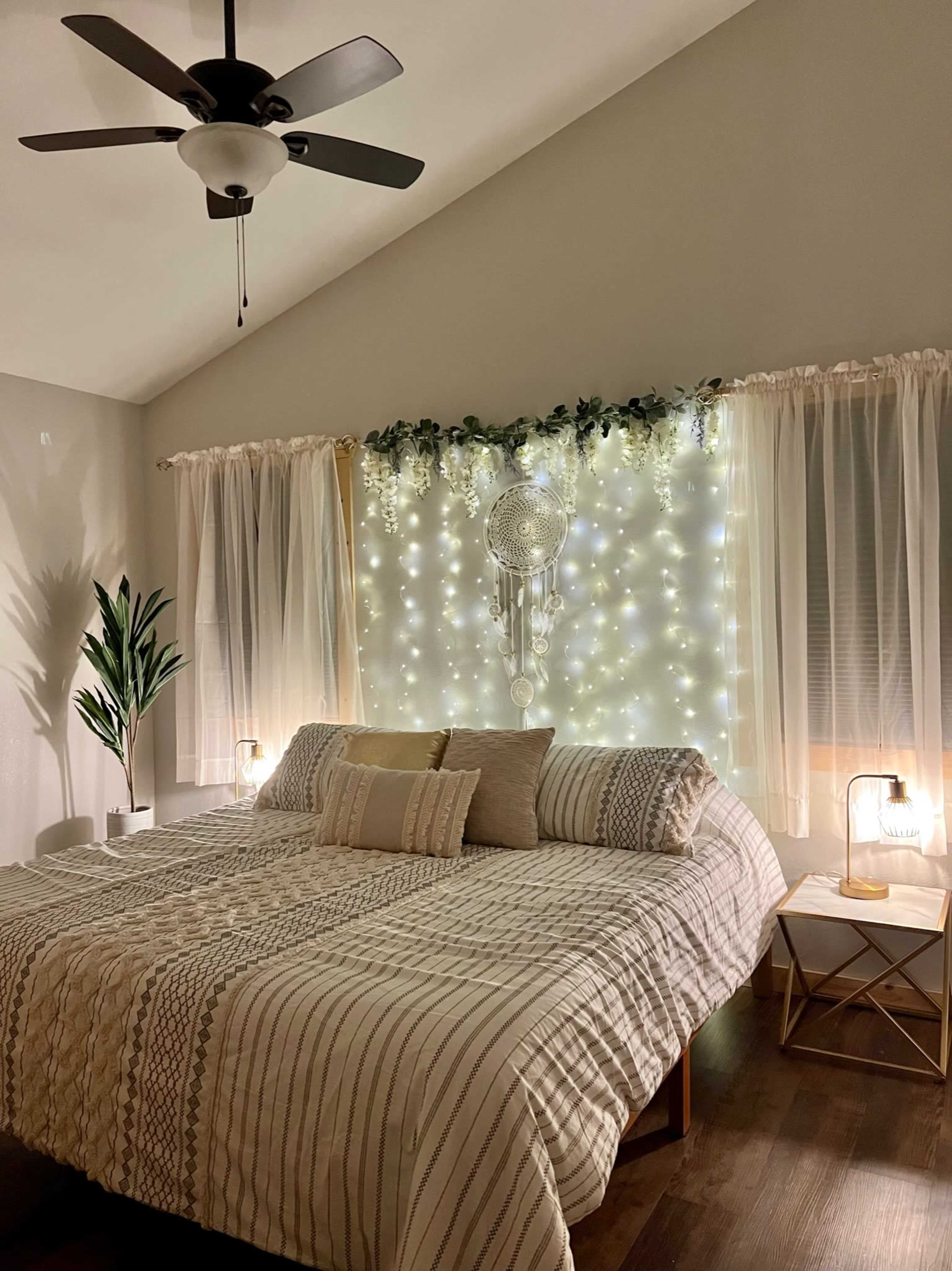 A bedroom with a large bed covered in a patterned blanket, a decorative wall adorned with fairy lights and a dreamcatcher, and two bedside lamps illuminating the space.