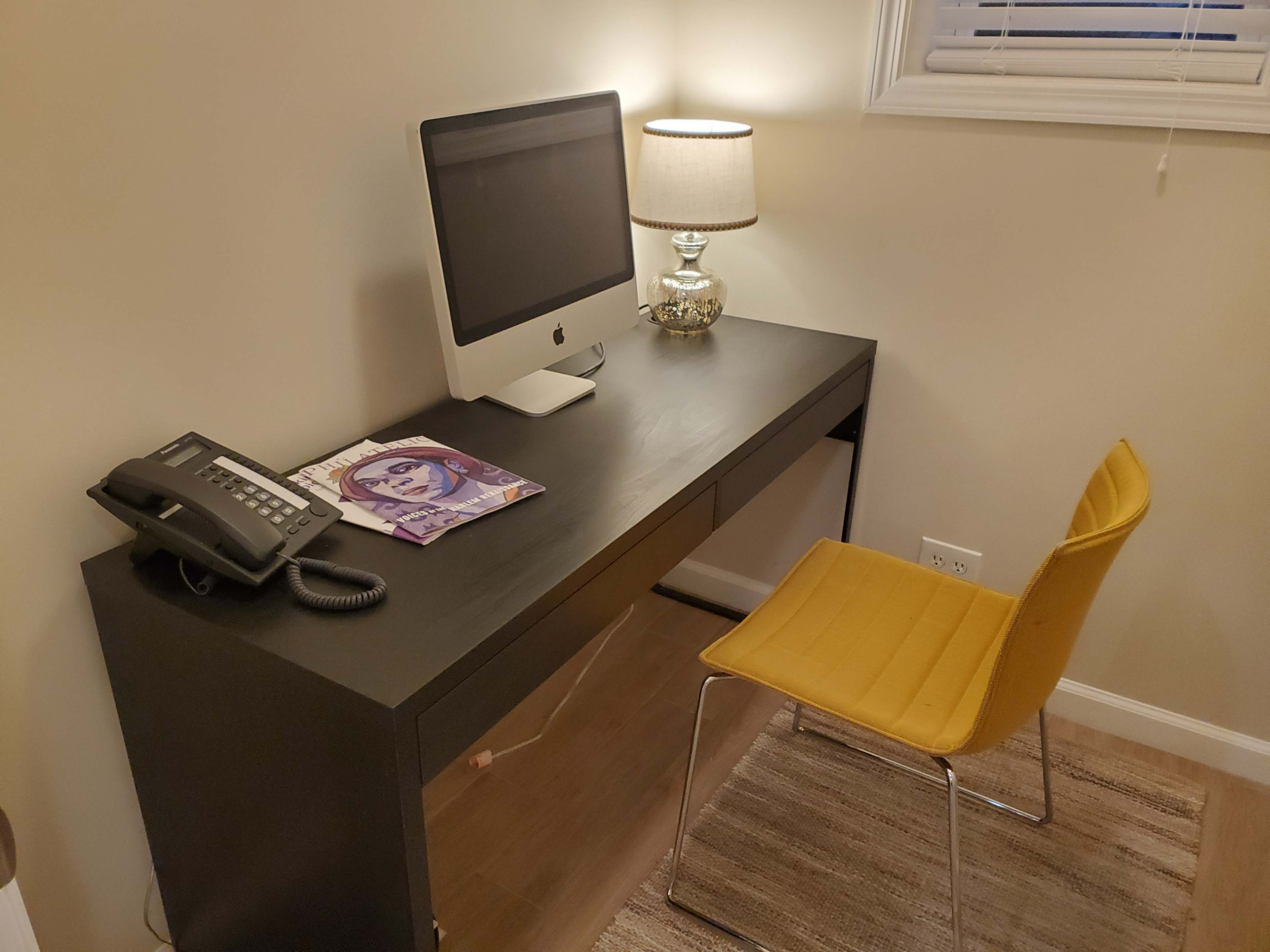 A black desk with a computer, a phone, a magazine, a yellow chair, and a lamp is positioned in a well-lit room.
