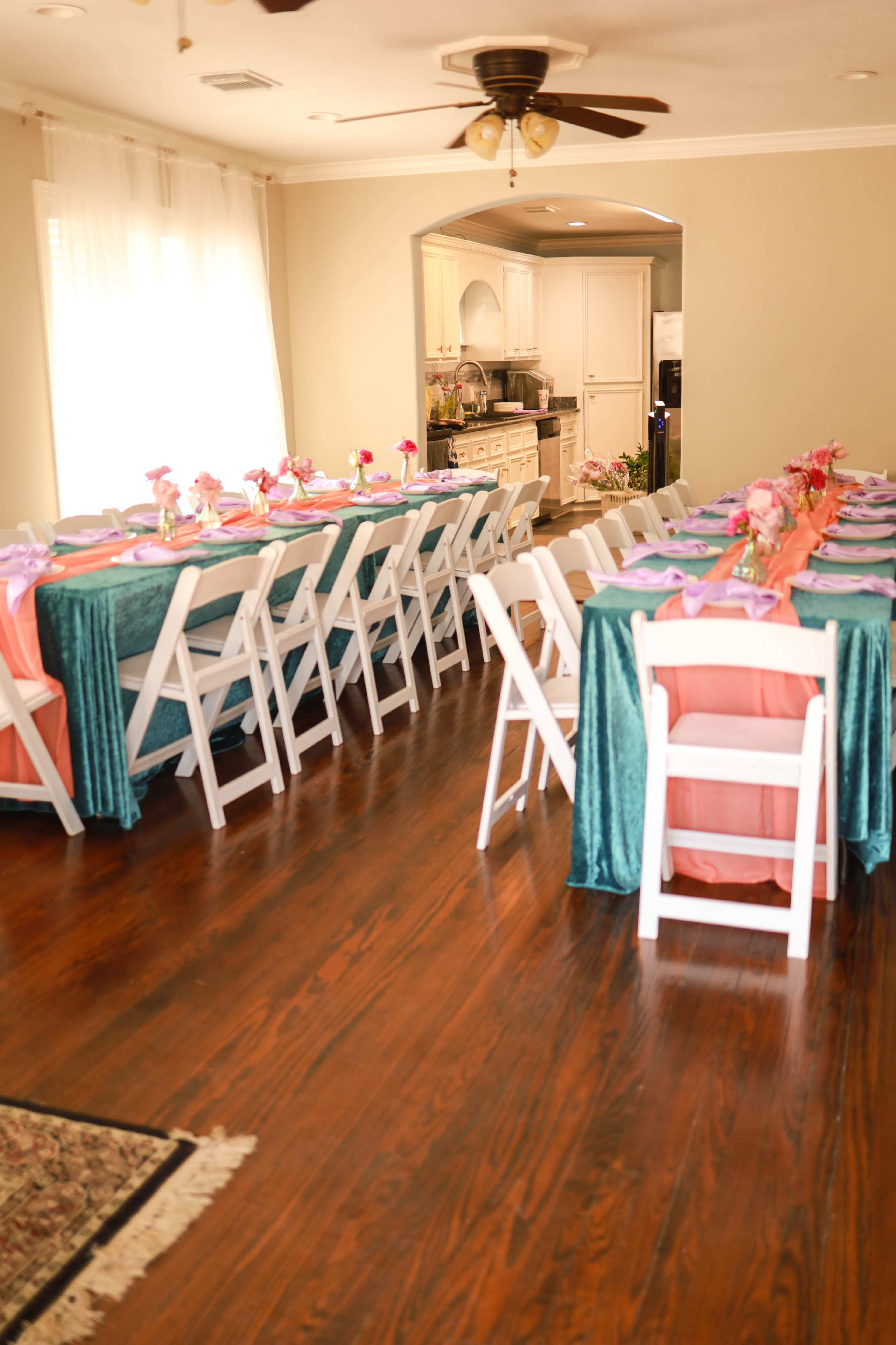 A dining area features multiple tables with teal and coral tablecloths set for a gathering, surrounded by white folding chairs.