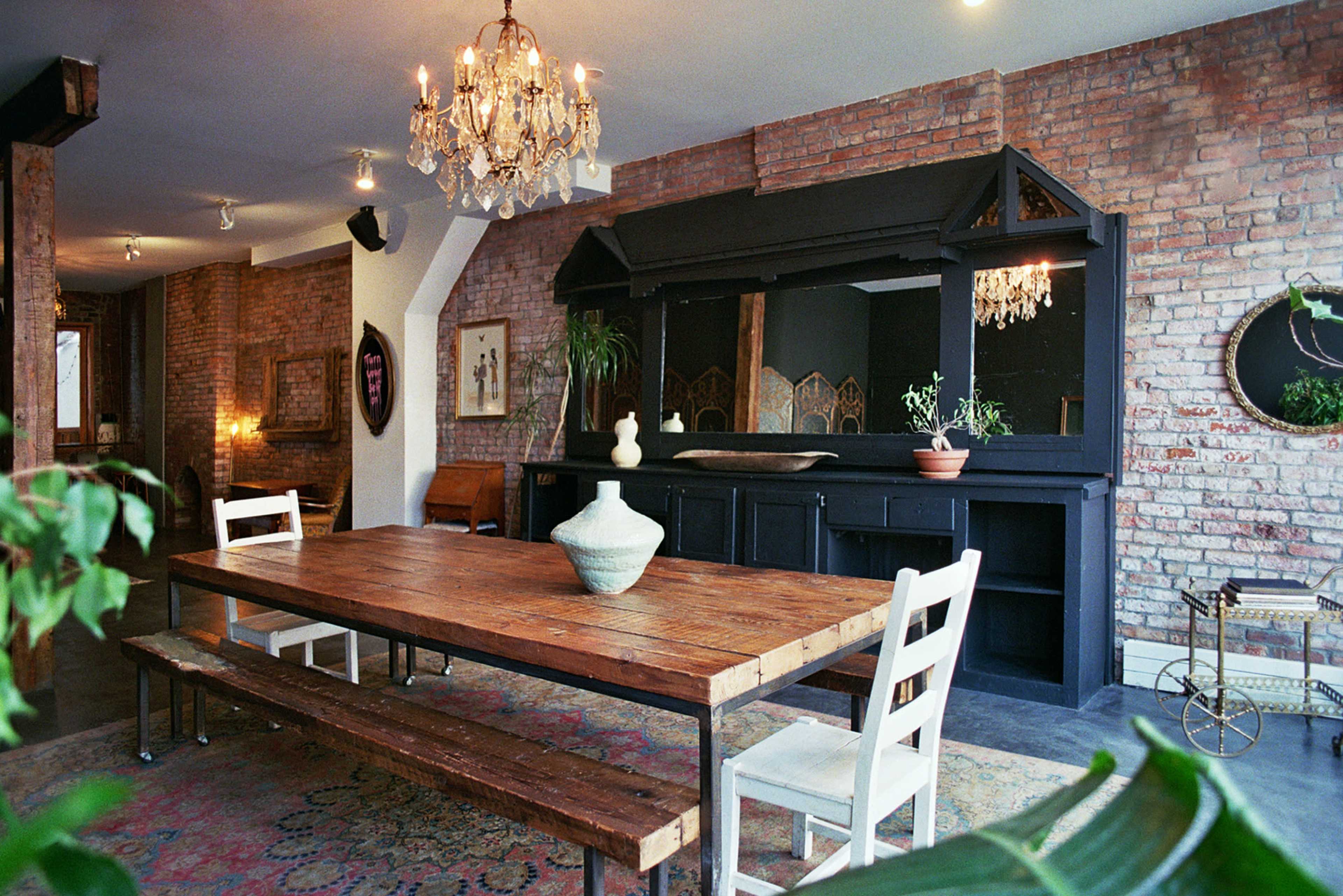 The image shows a rustic dining area featuring a large wooden table, white chairs, and a decorative black wooden sideboard against a brick wall.