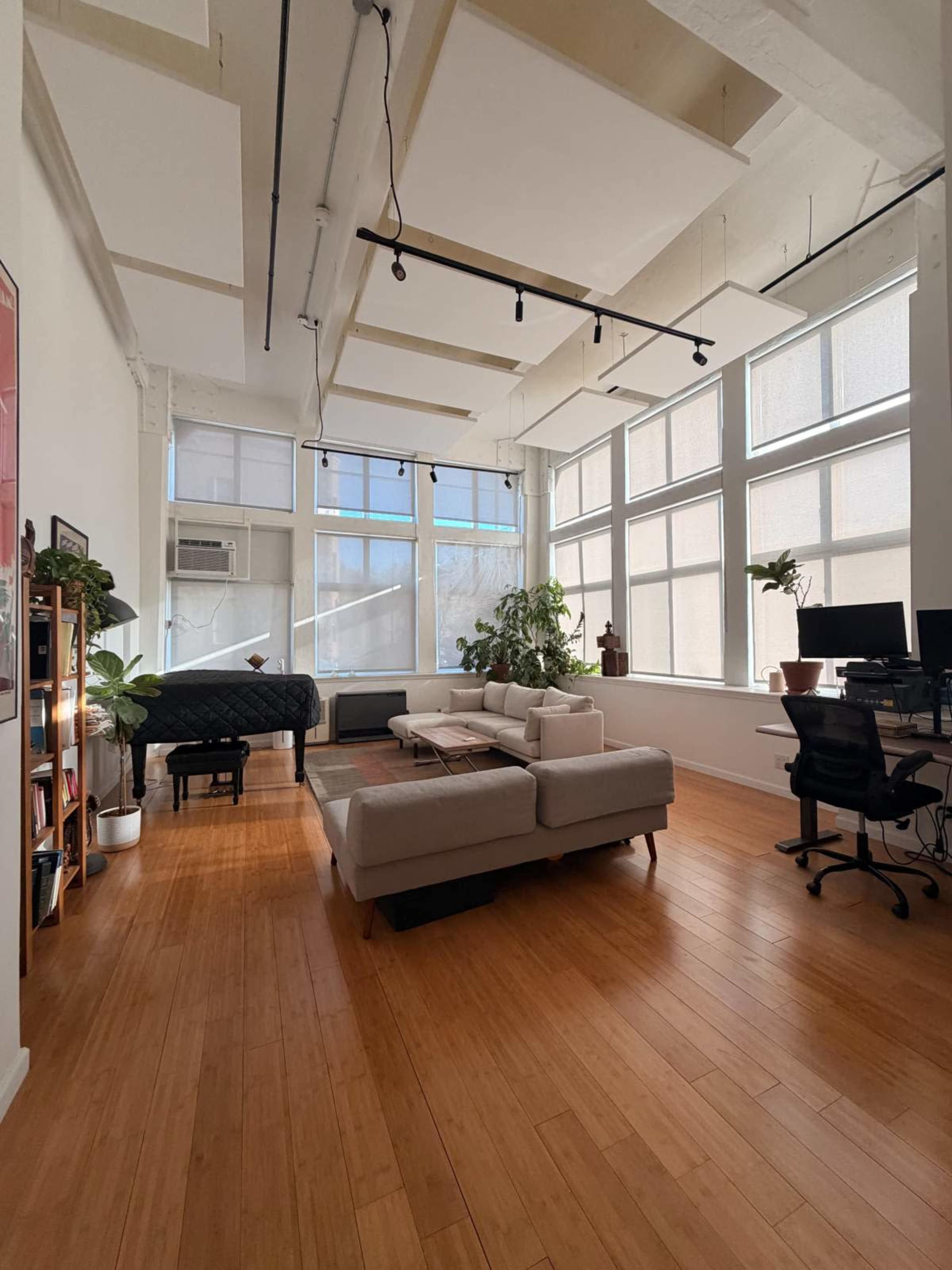 The image shows a spacious and bright room with large windows, a piano in one corner, a seating area with a sofa, and a workspace with a desk and computer.