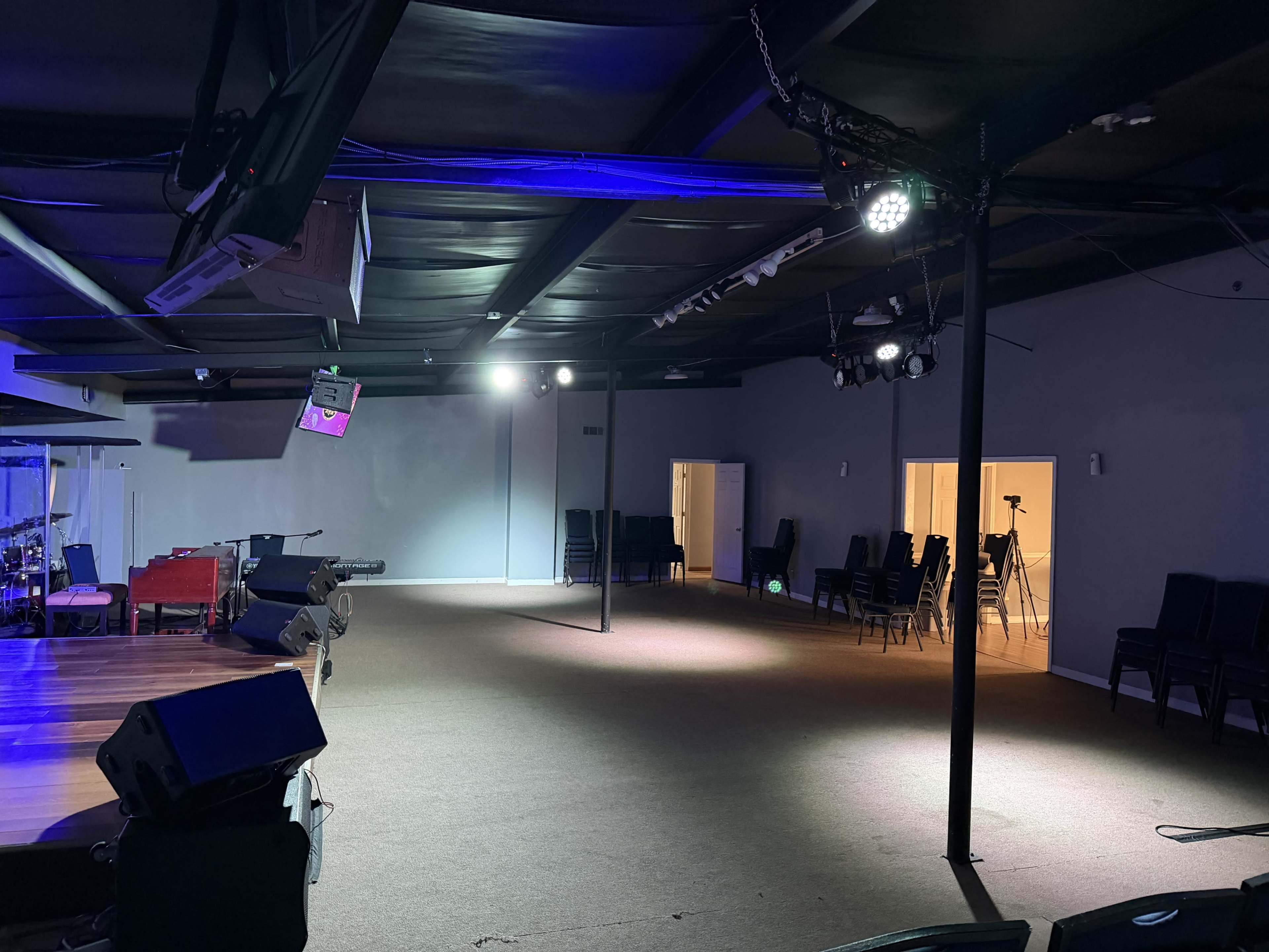 The image shows a large, empty indoor space with a stage area and rows of black chairs arranged against the walls.