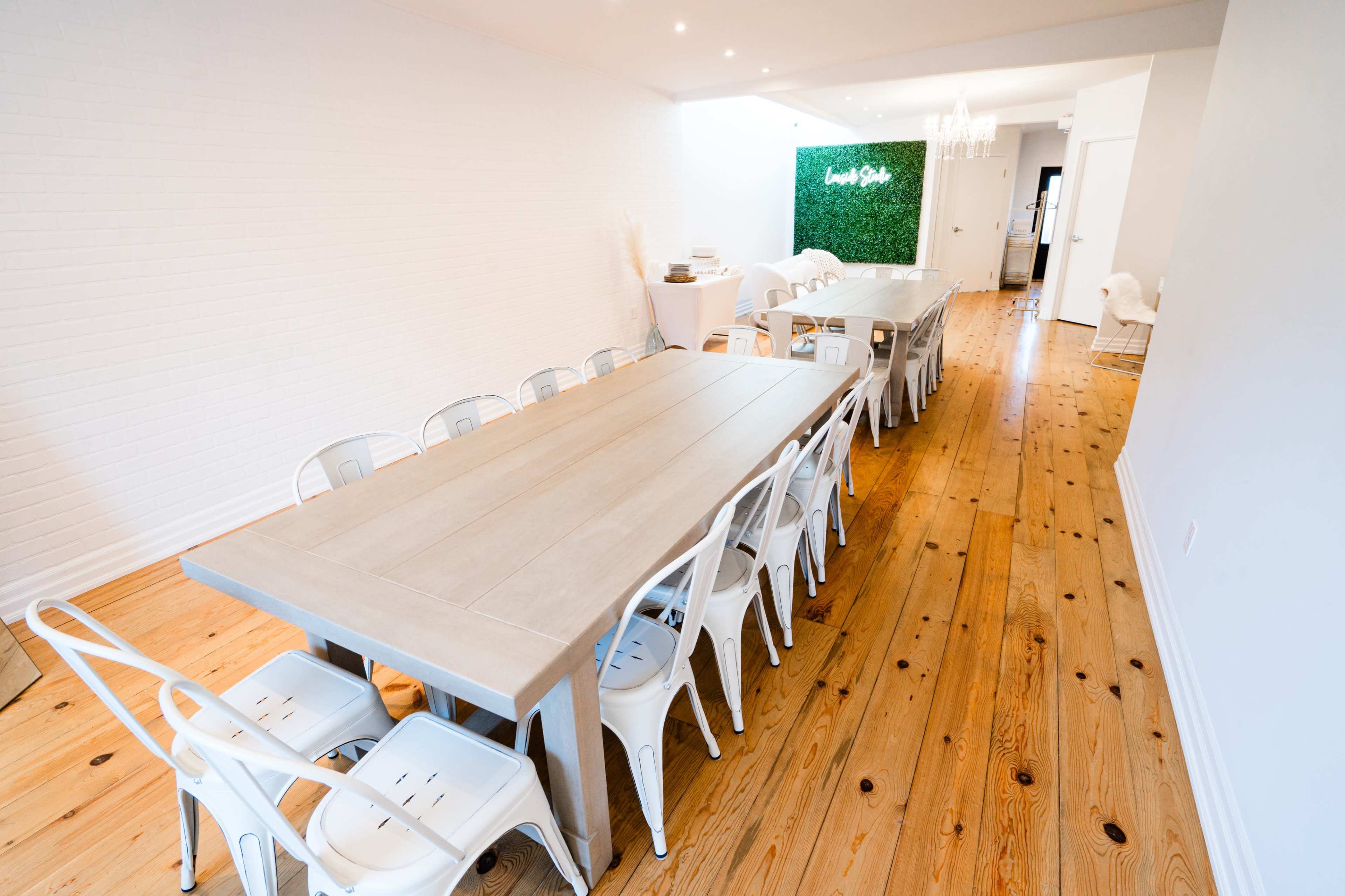 A long rectangular table with white chairs is set up in a spacious room with wooden floors and green wall decor at one end.