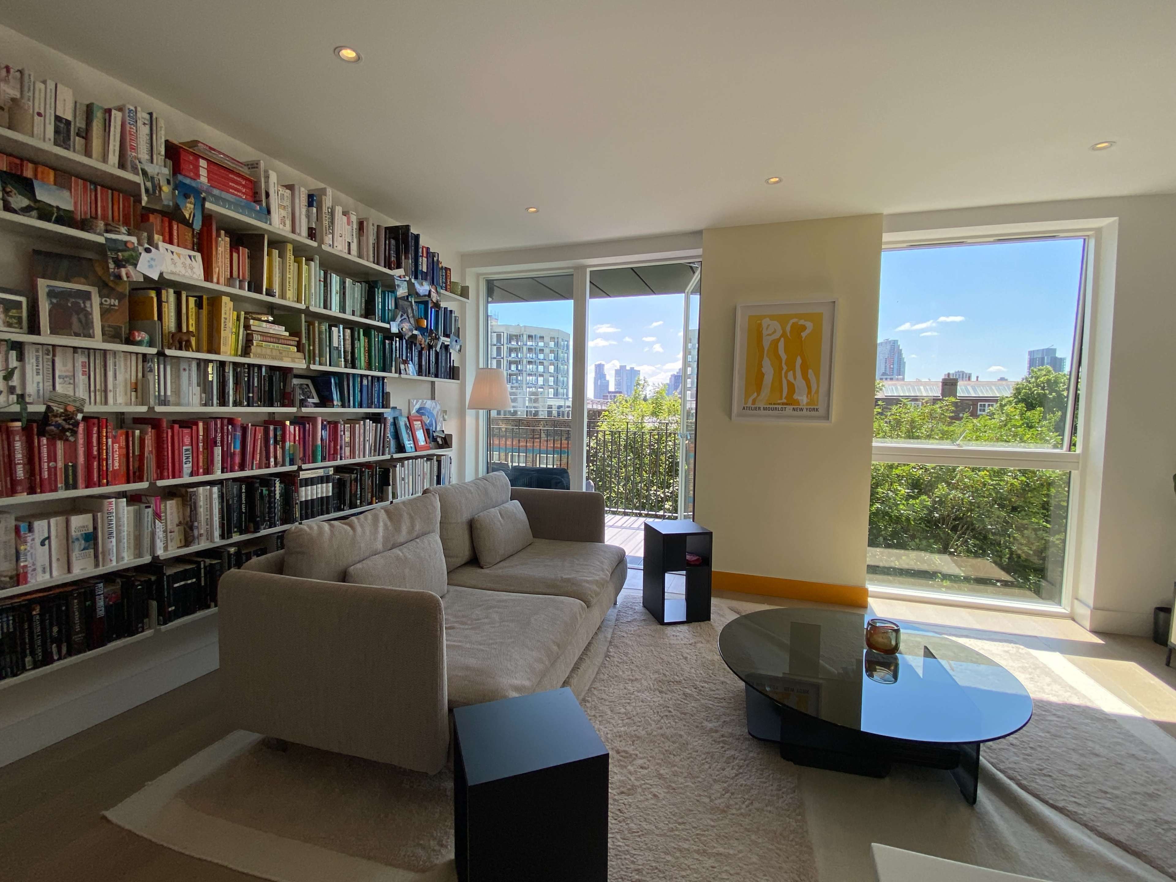 A modern living room features a large bookshelf filled with colorful books, a beige sofa, a glass coffee table, and a view of the cityscape through a large window.