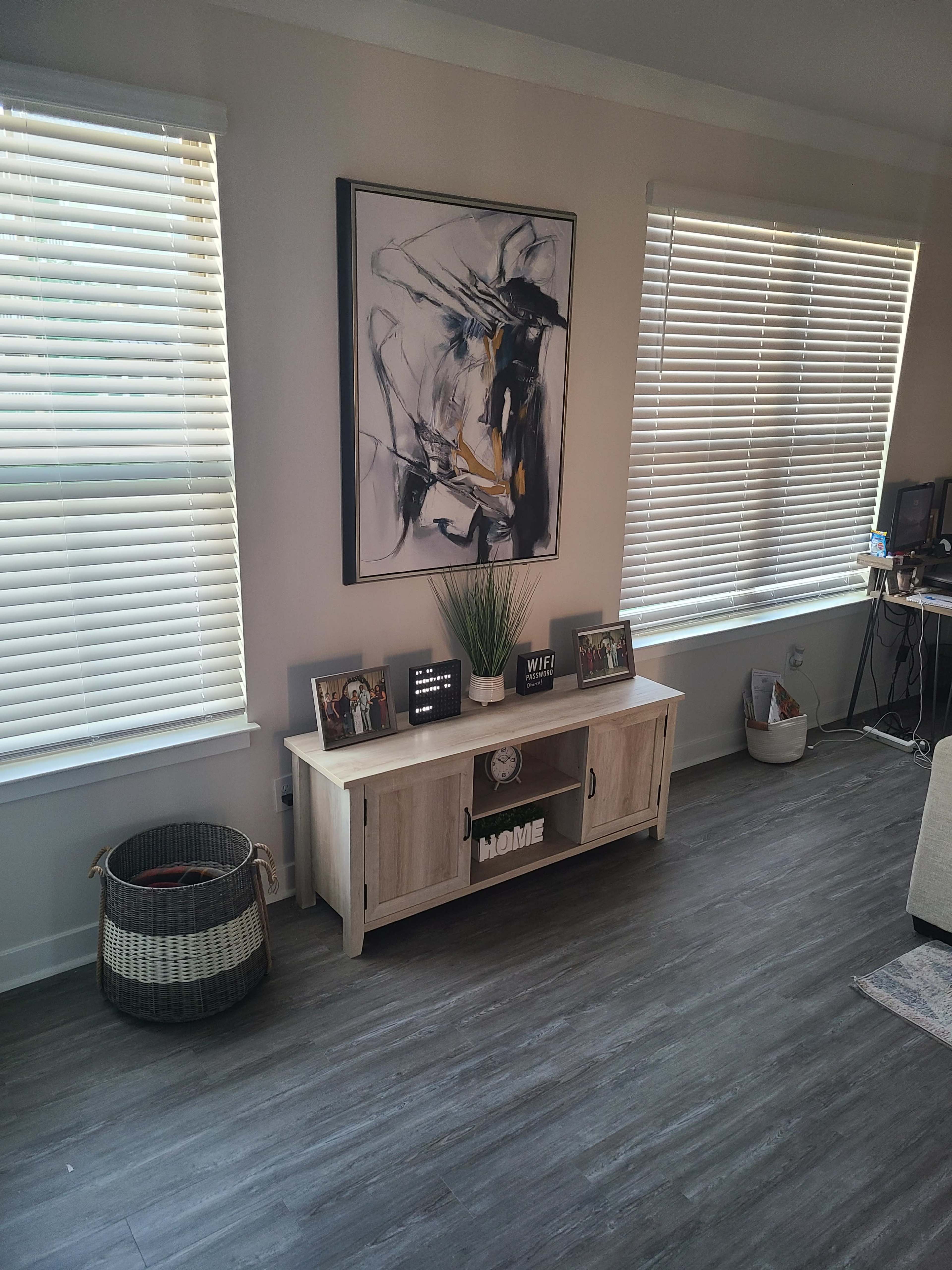 A living room with a modern wooden entertainment center beneath an abstract painting, framed photos, and a decorative plant, with two windows featuring blinds allowing light to filter in.