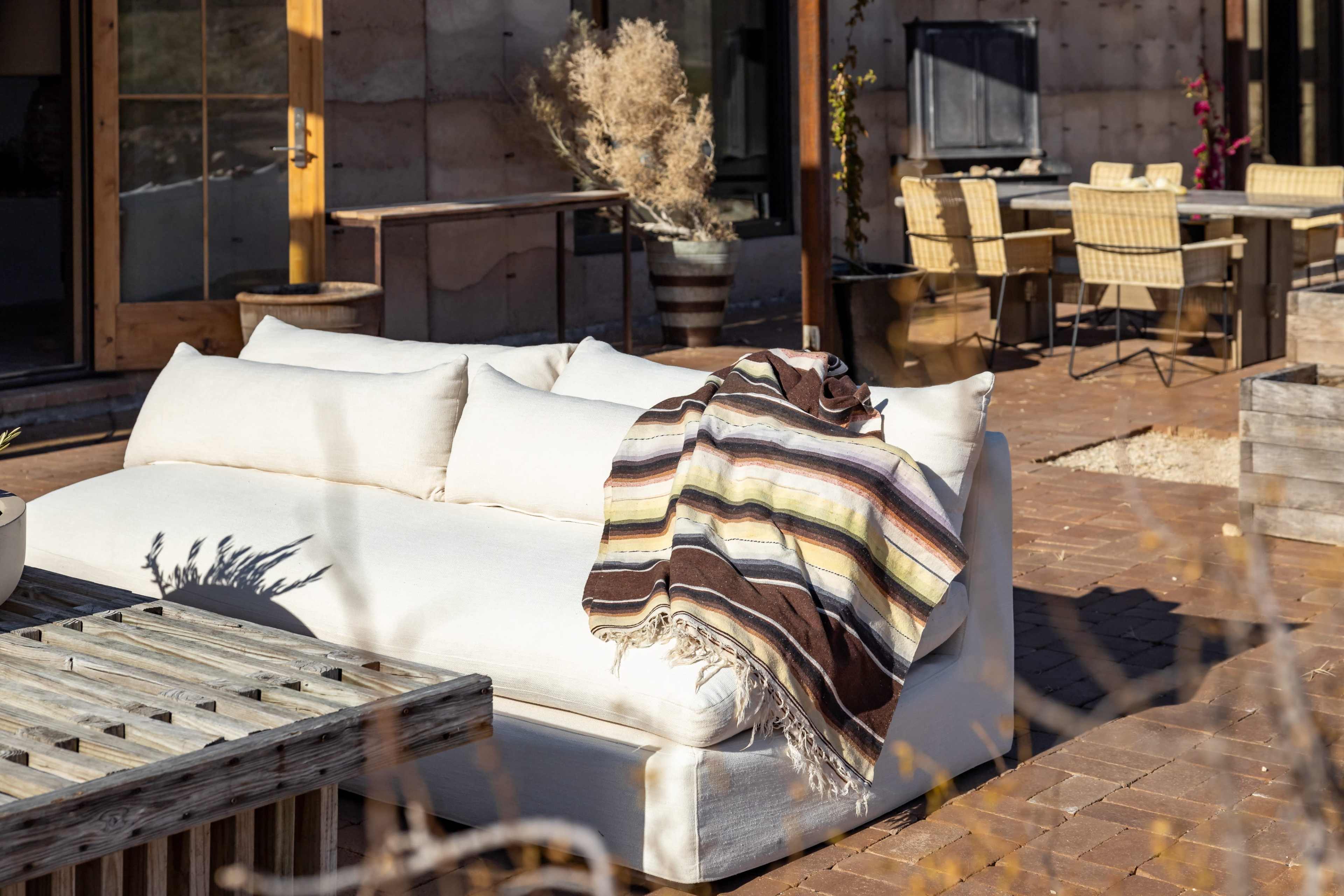 A white outdoor sofa with a striped blanket rests on a patio with wooden furniture and potted plants.