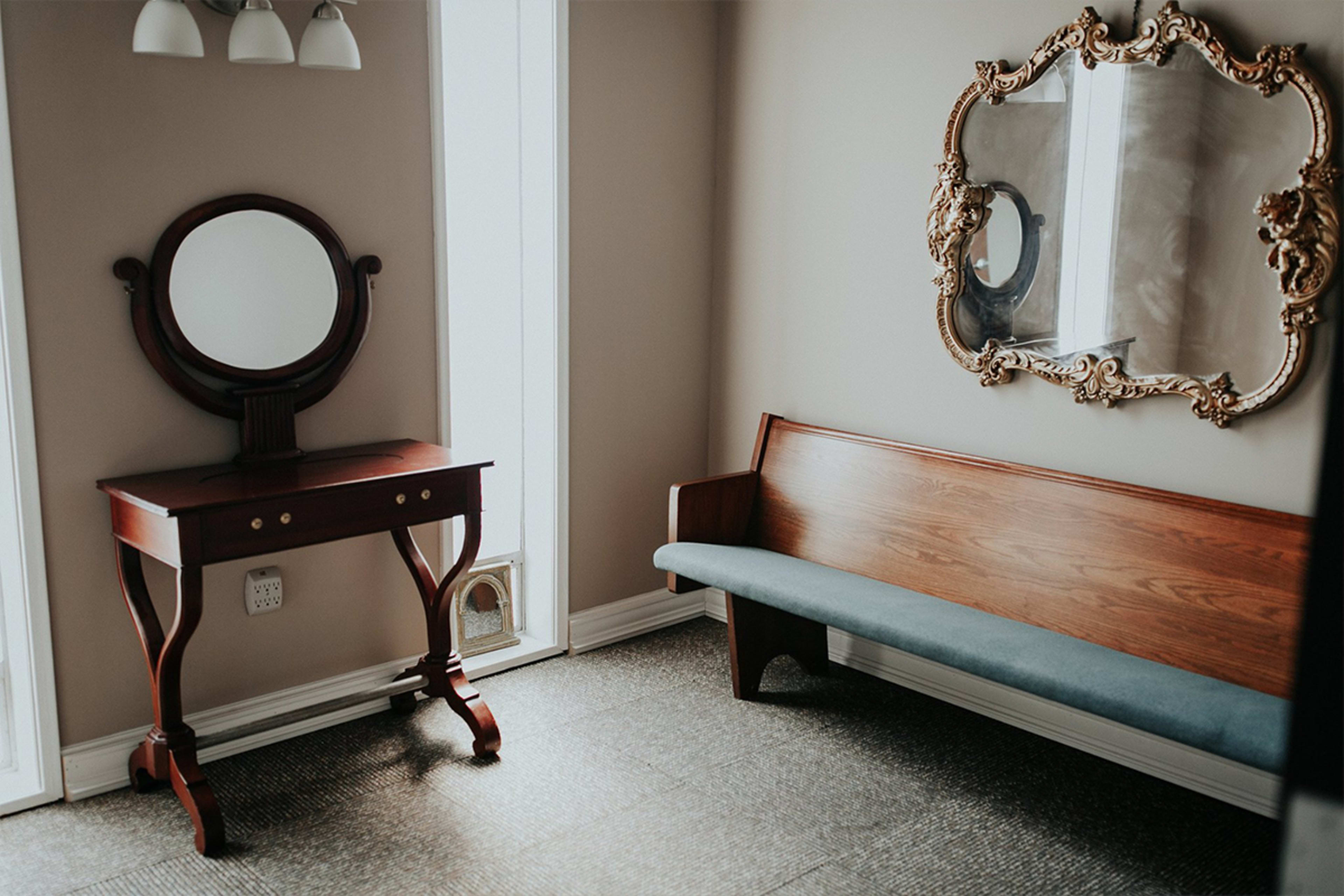 A simple interior space with a wooden bench, a decorative mirror, and a small vanity table against a beige wall.
