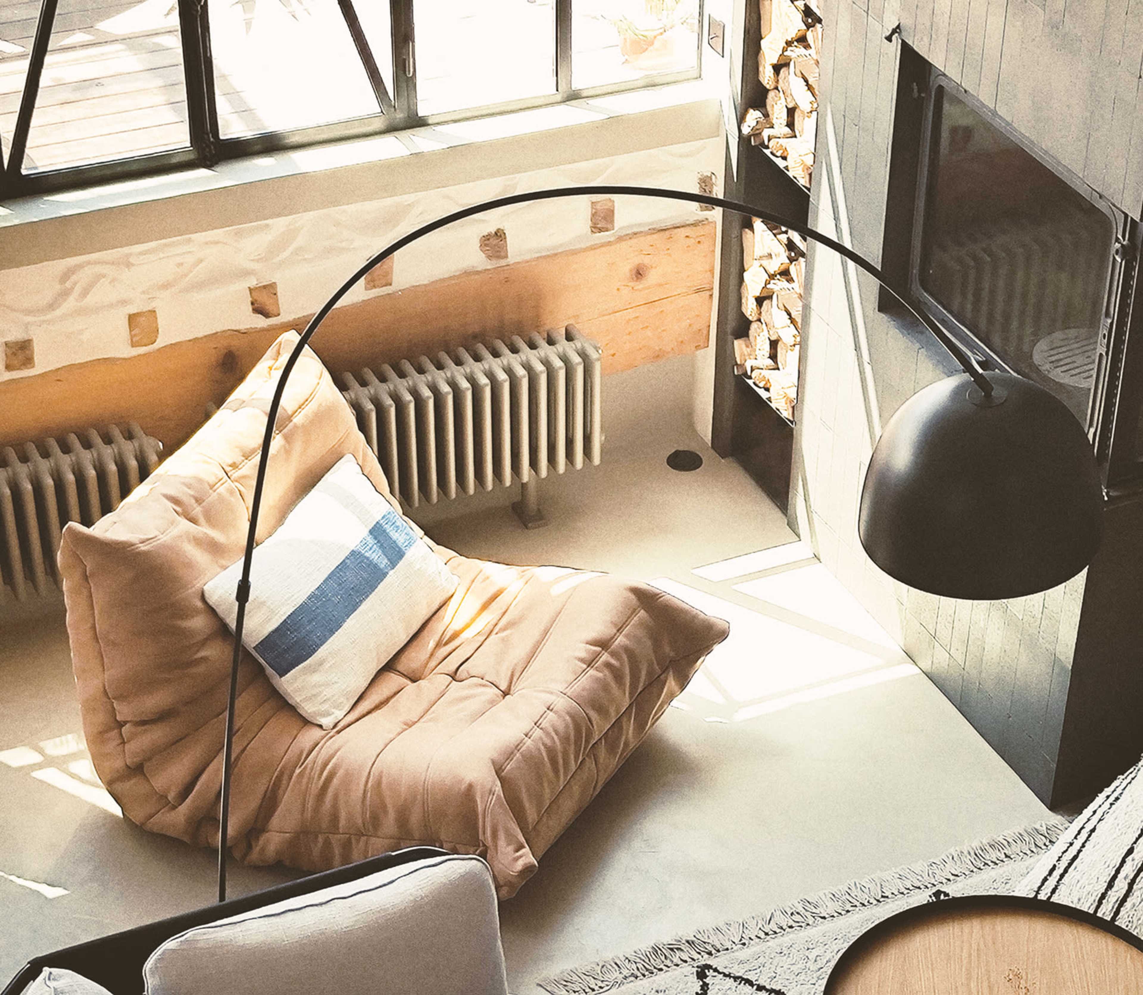 A large, cushioned chair with a blue and white striped pillow sits near a modern fireplace, illuminated by natural light streaming through large windows.