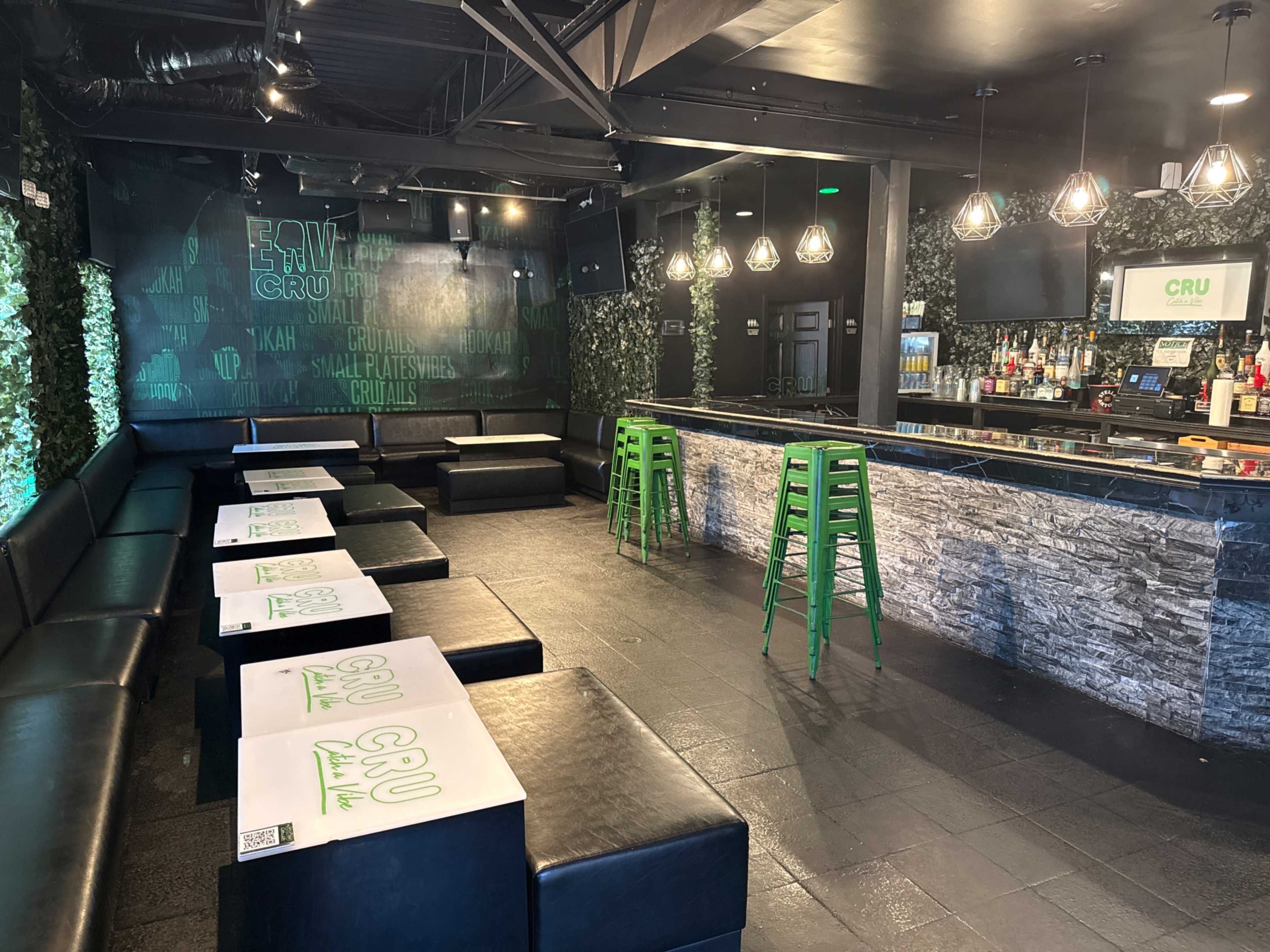 The image shows a modern bar area with dark furnishings, a stone bar, and green accents, including stools and wall decor.