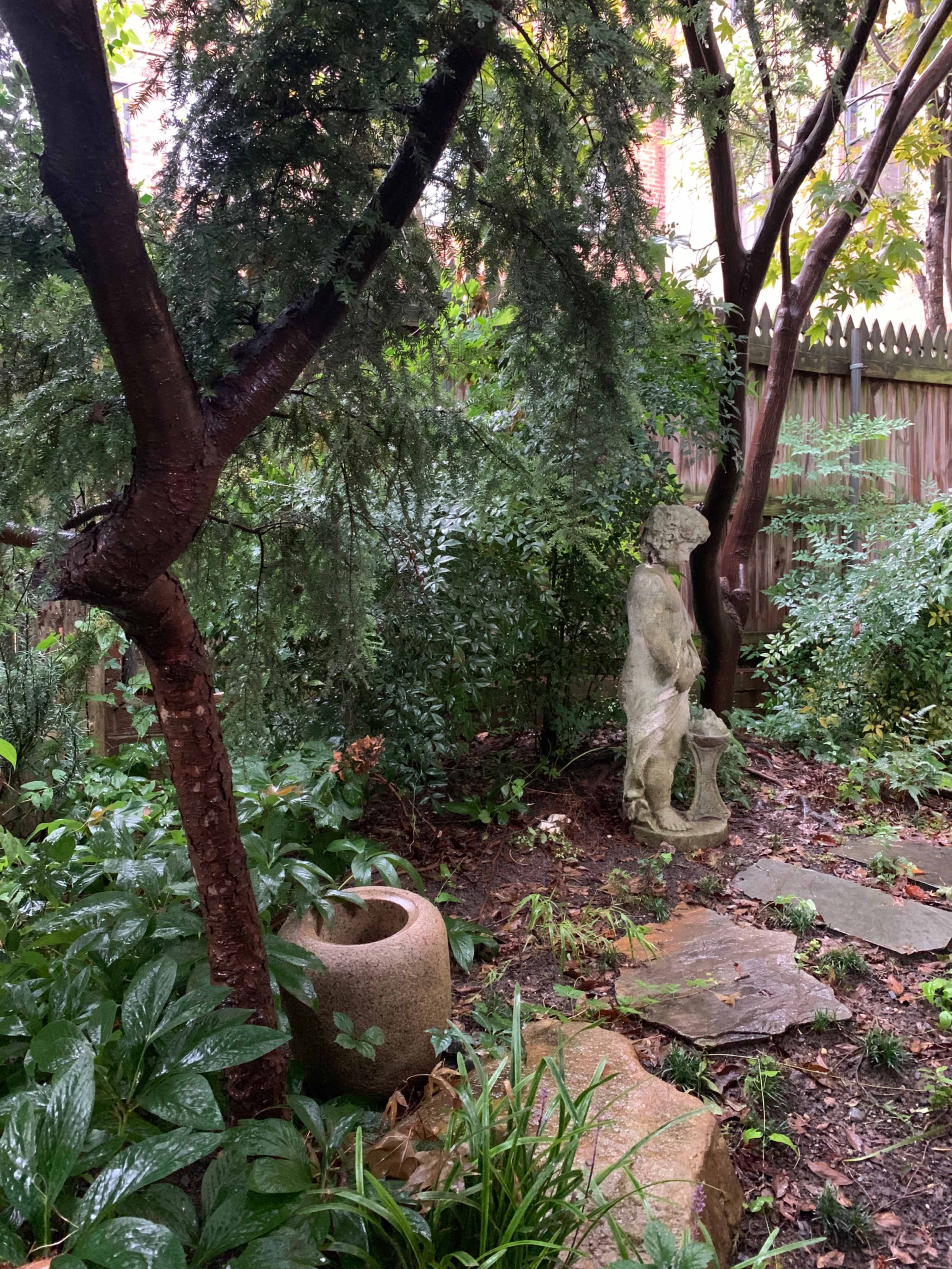 A serene garden featuring a stone sculpture, a circular stone planter, and lush greenery surrounded by trees.
