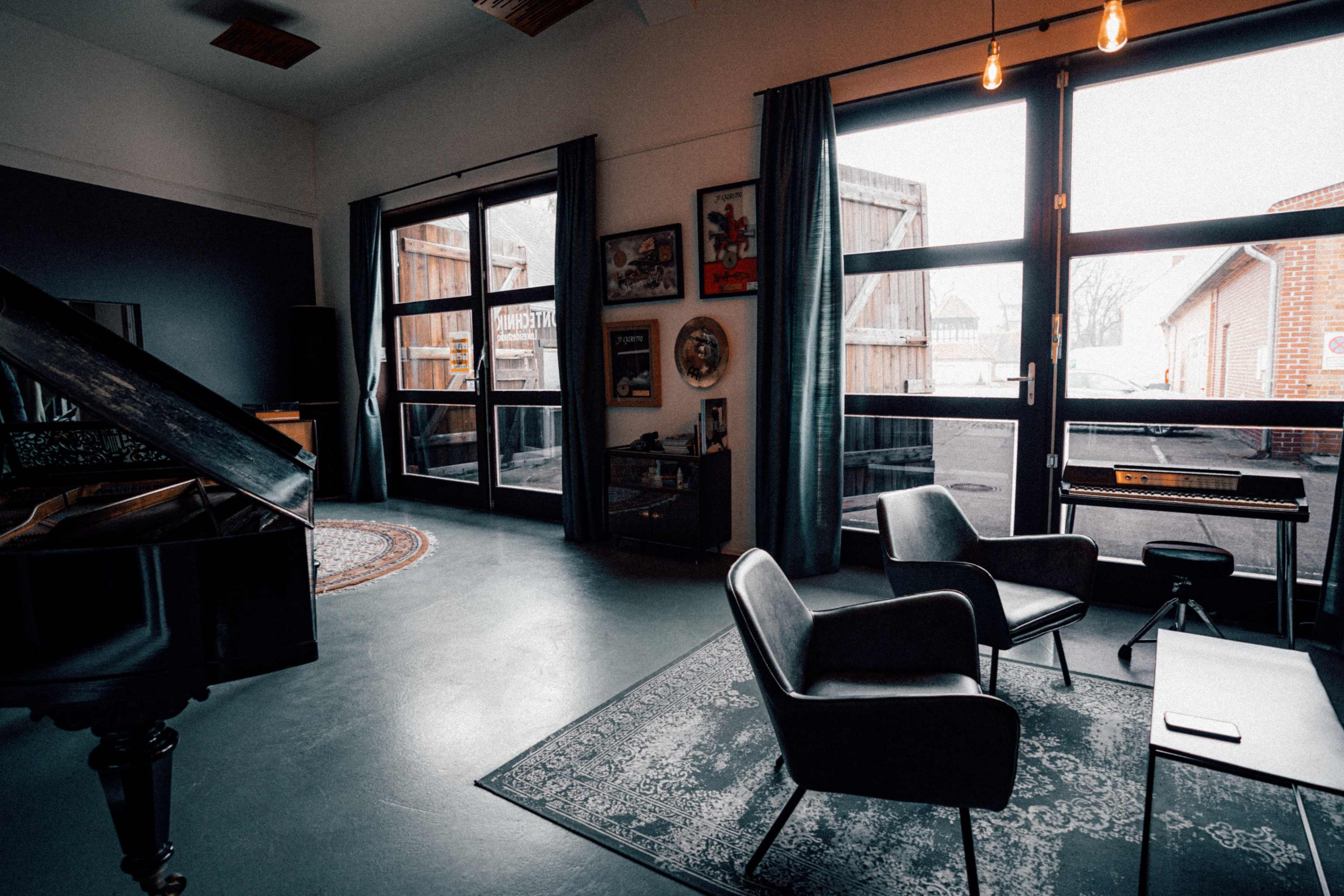 A spacious indoor room features a grand piano, two chairs, a small table, and large windows that overlook an outdoor area.