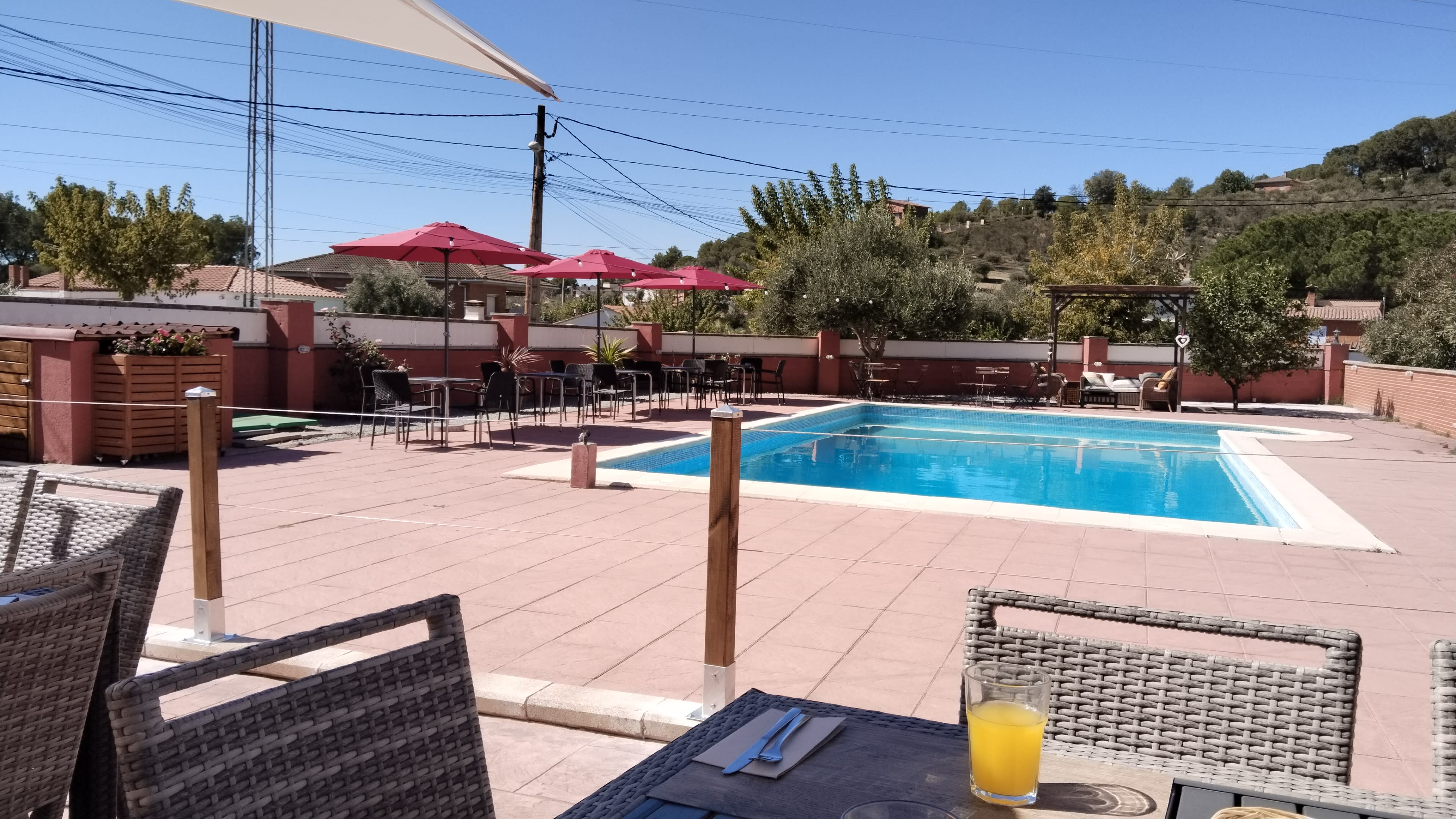 The scene shows a swimming pool surrounded by patio furniture and shaded by umbrellas, with a table set for dining in the foreground.