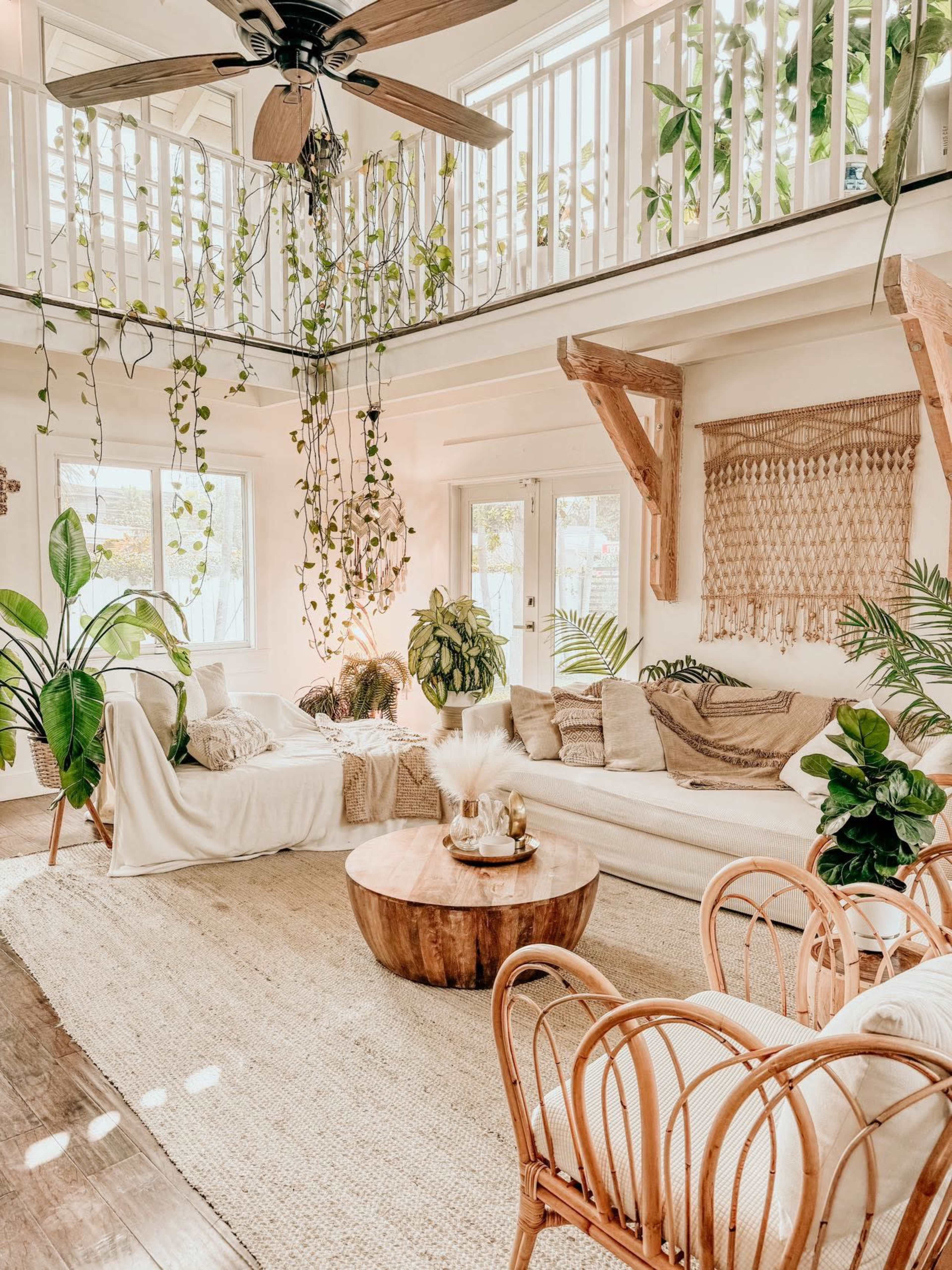 The image shows a bright, open living room with large windows, a wooden coffee table, a sectional sofa, potted plants, and hanging greenery.