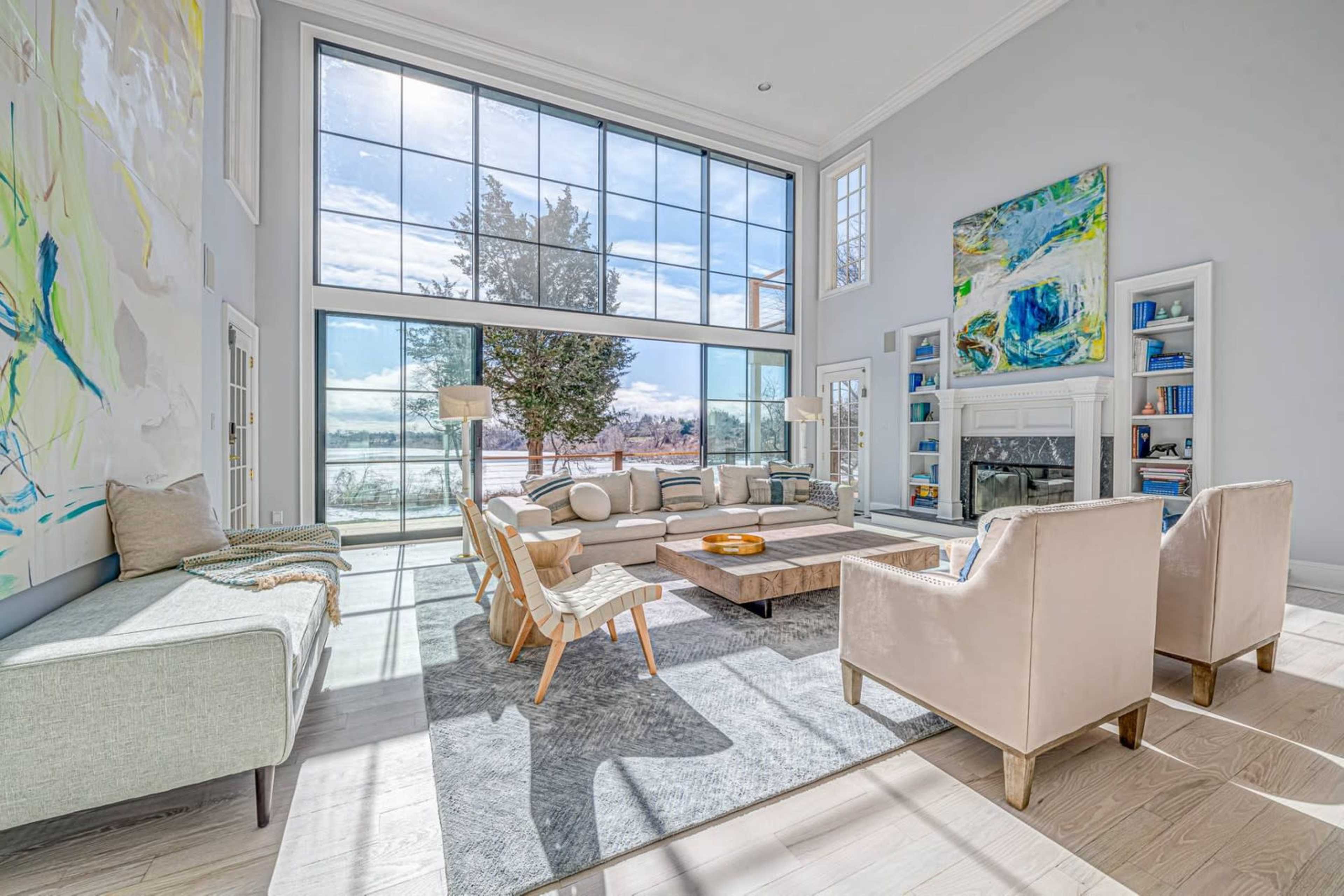 The living room features large windows that provide a view of the outdoors, with a minimalist furniture arrangement including a sectional sofa, armchairs, and a coffee table set on a rug.