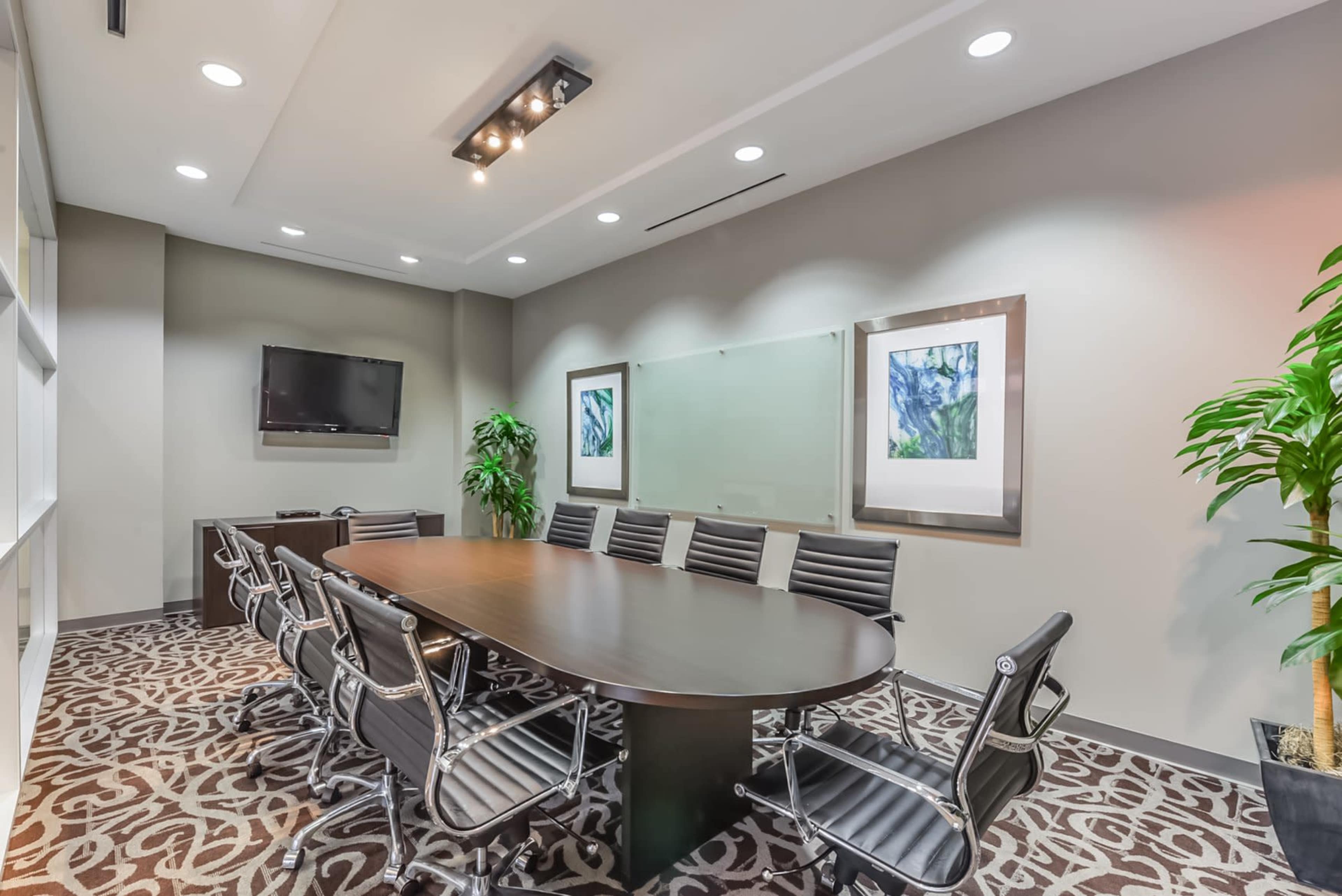 A modern conference room features a long table surrounded by black chairs, with two framed artworks and a television on the wall.