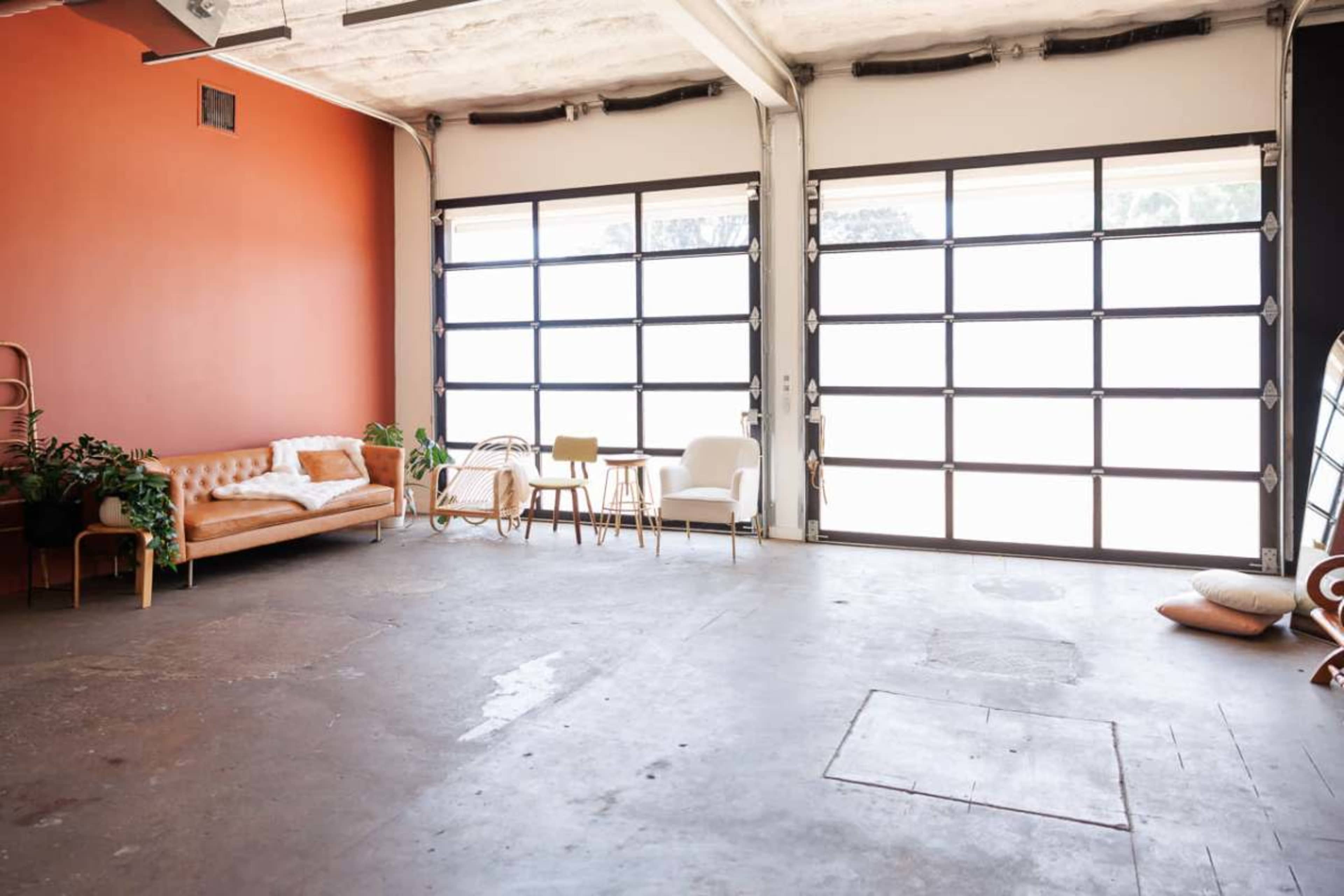 The image shows an industrial-style interior space with a brown leather sofa, a small round table, two chairs, and large glass garage doors allowing natural light.
