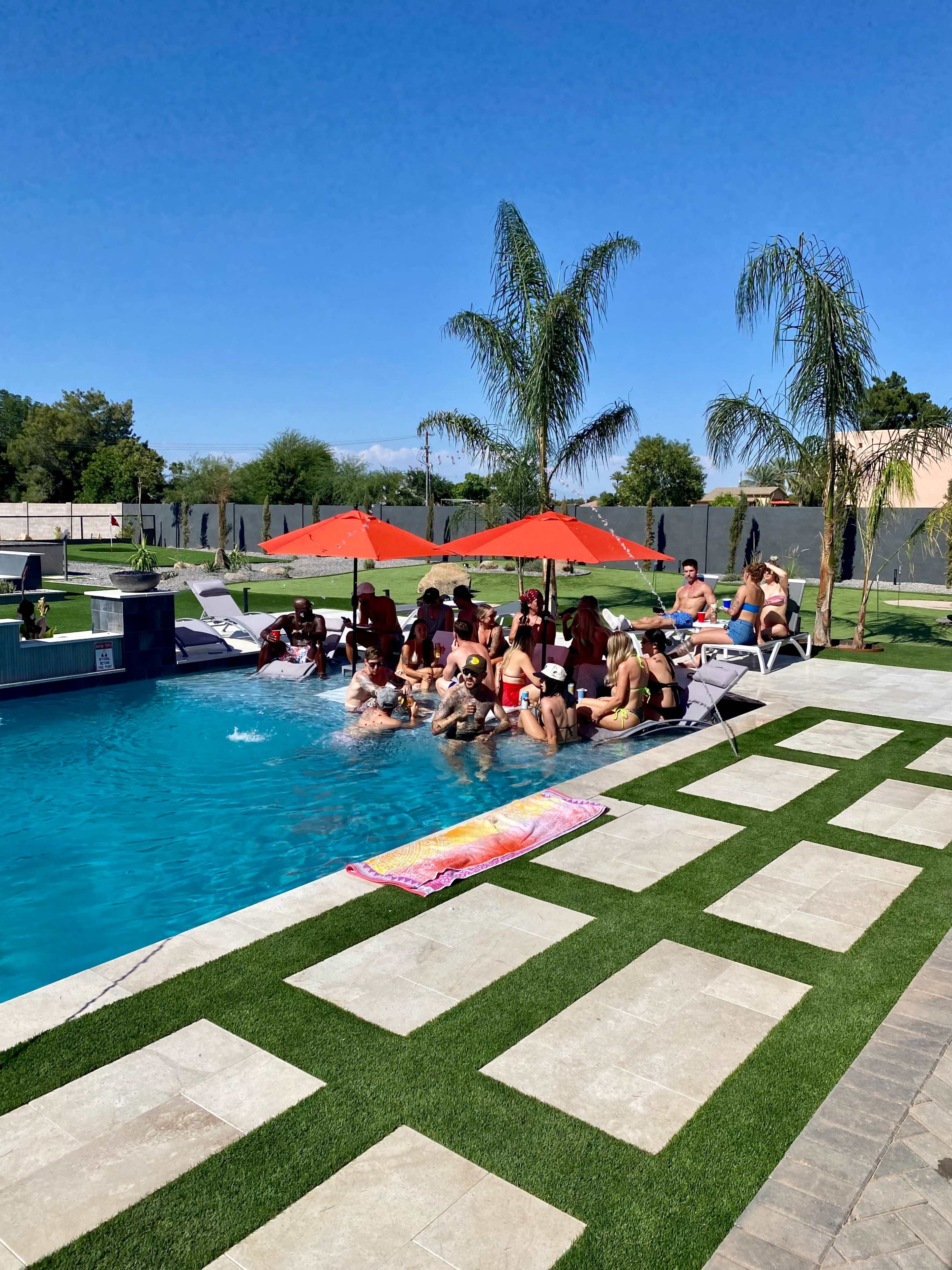 A group of people is gathered around a swimming pool, sitting on lounge chairs and in the water, under red umbrellas on a sunny day.
