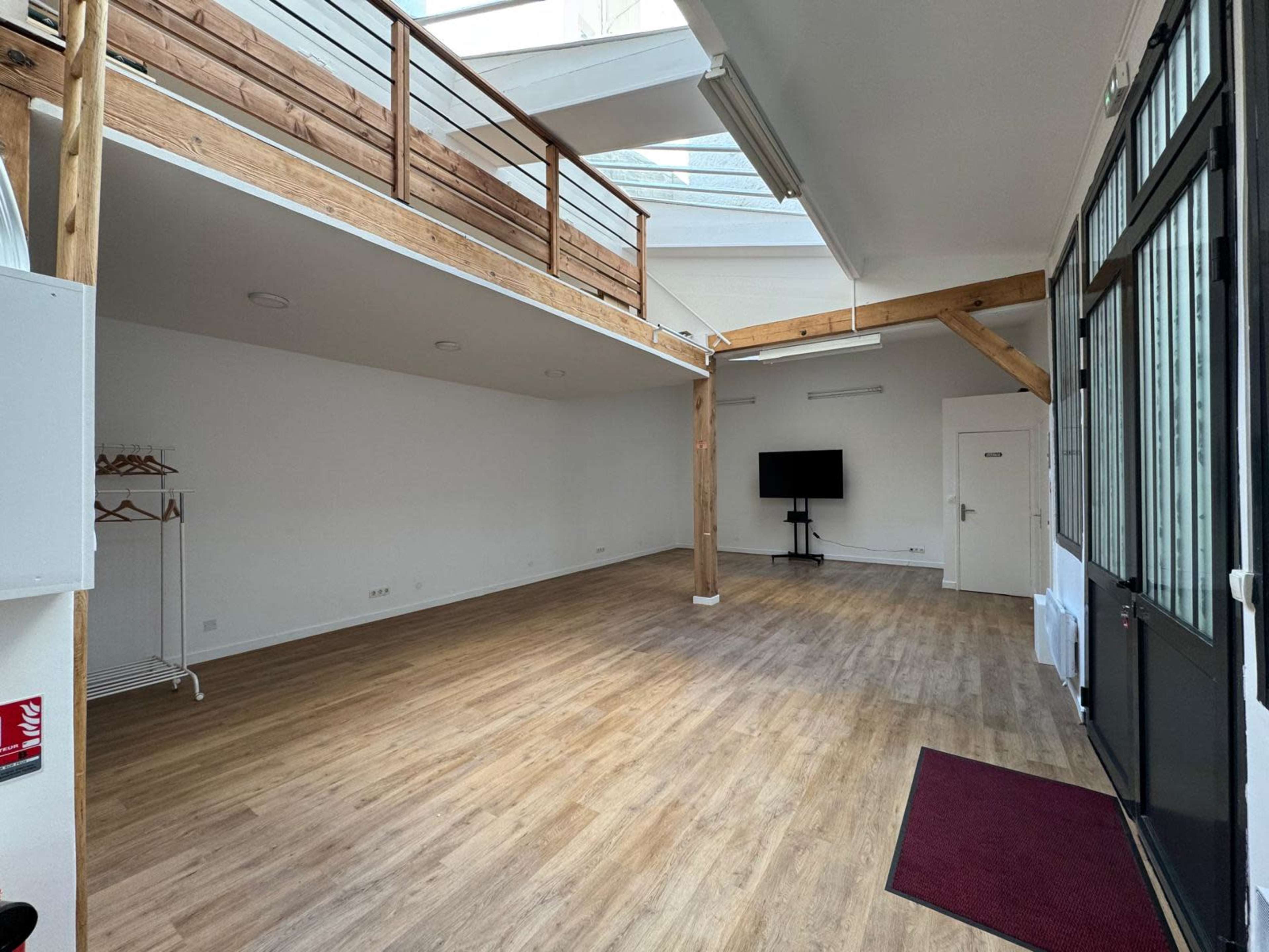 The image shows an empty, spacious interior with wooden flooring, large windows, and a loft area supported by wooden beams.