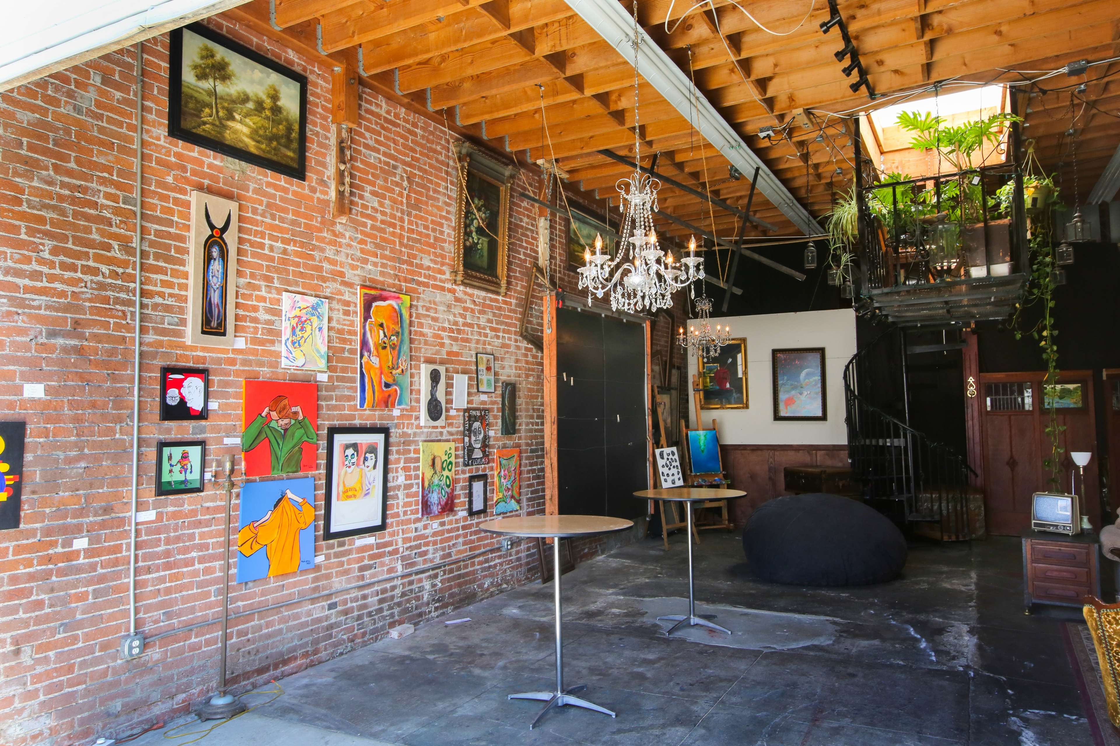 The image shows an industrial-style interior featuring exposed brick walls, an assortment of colorful artwork, two small tables, and a chandelier, with a staircase leading to a lofted area adorned with plants.
