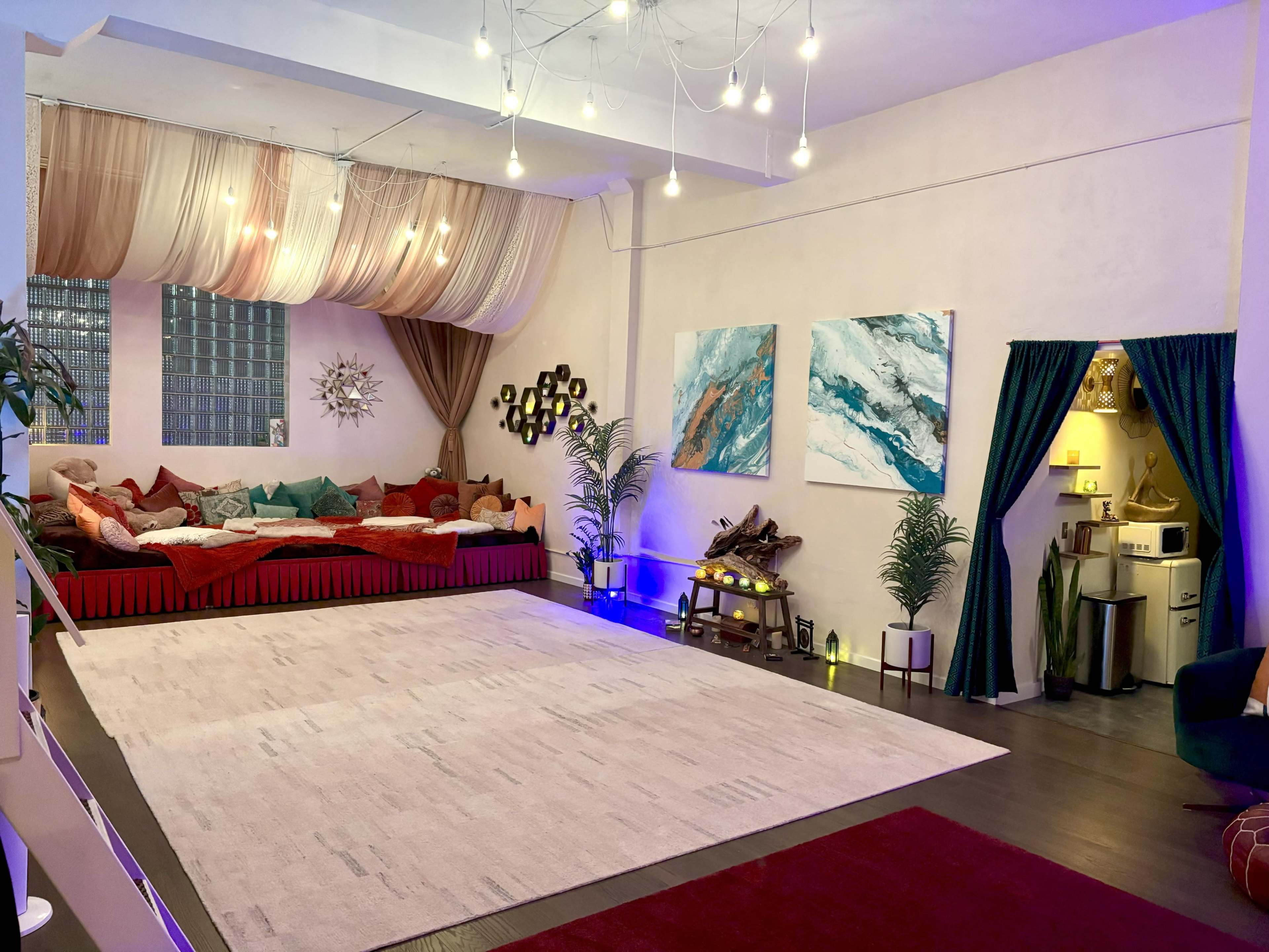 The spacious room features a large area rug, vibrant paintings on the walls, a cozy seating area with colorful cushions, and an entrance to a small kitchen space.