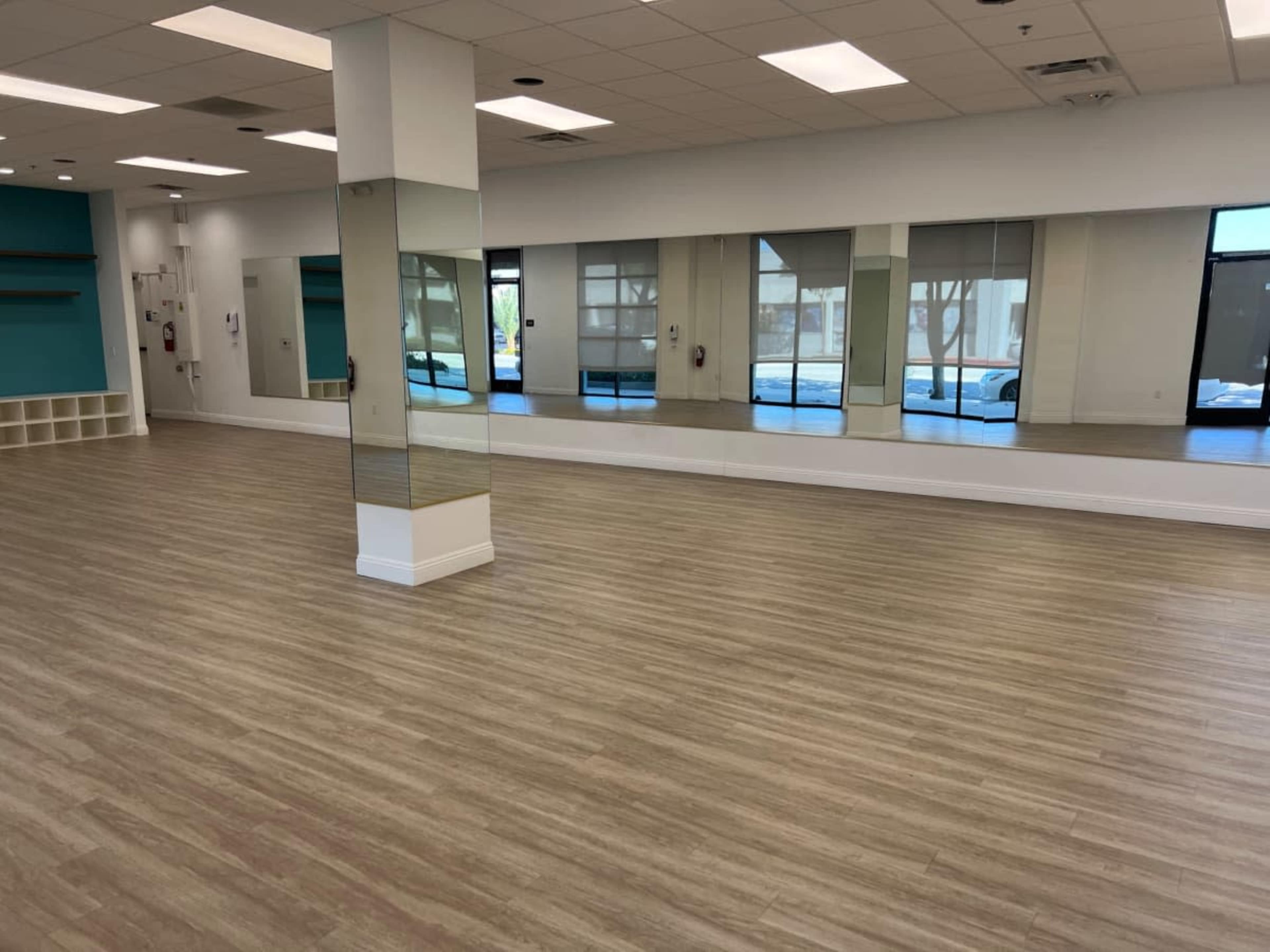 A spacious room with wooden flooring, mirrored walls, and large windows allowing natural light to enter.