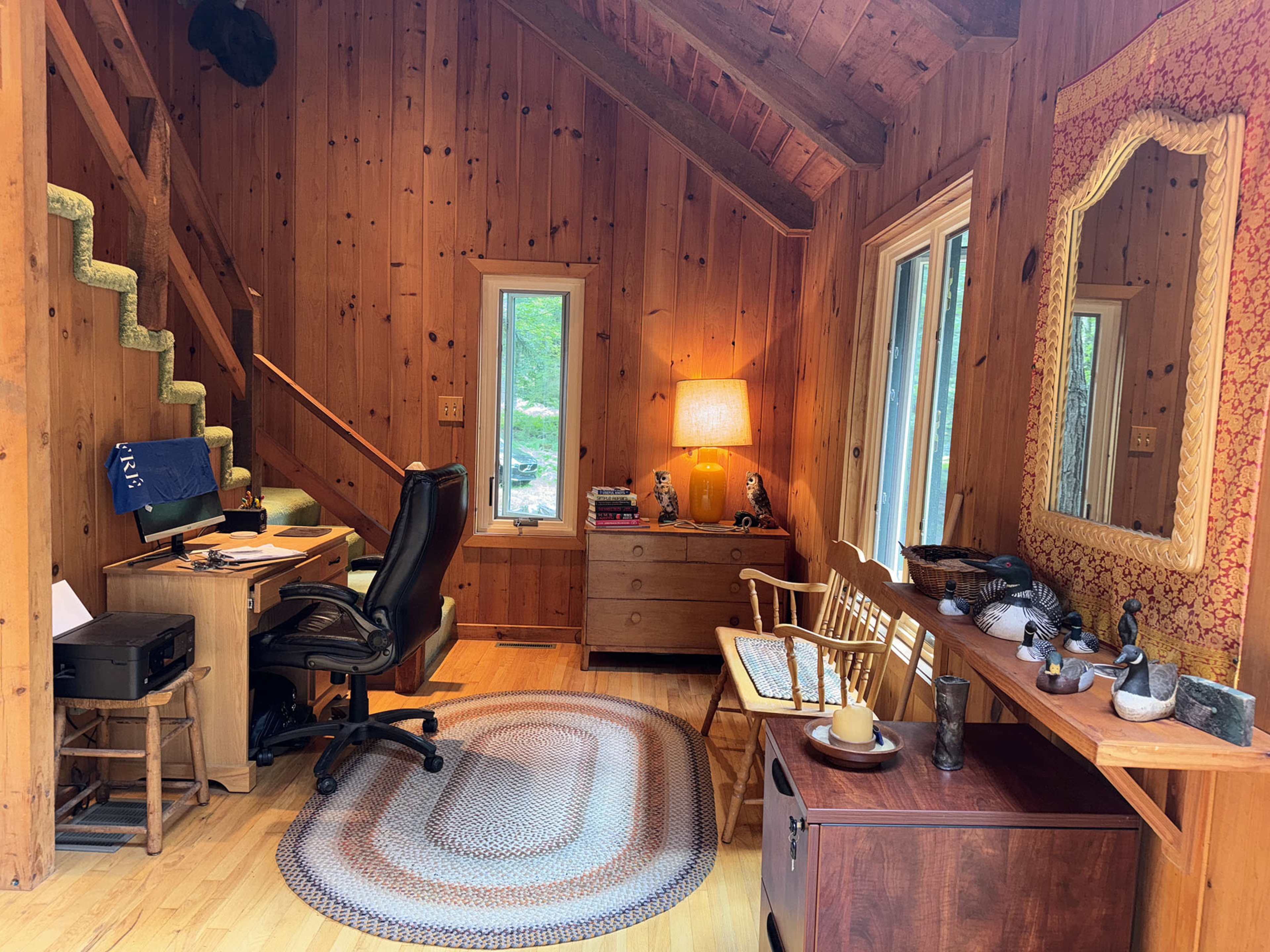 The image shows a cozy wood-paneled room featuring a desk with a chair, a decorative wall mirror, a small bench, and a staircase leading to an upper level.
