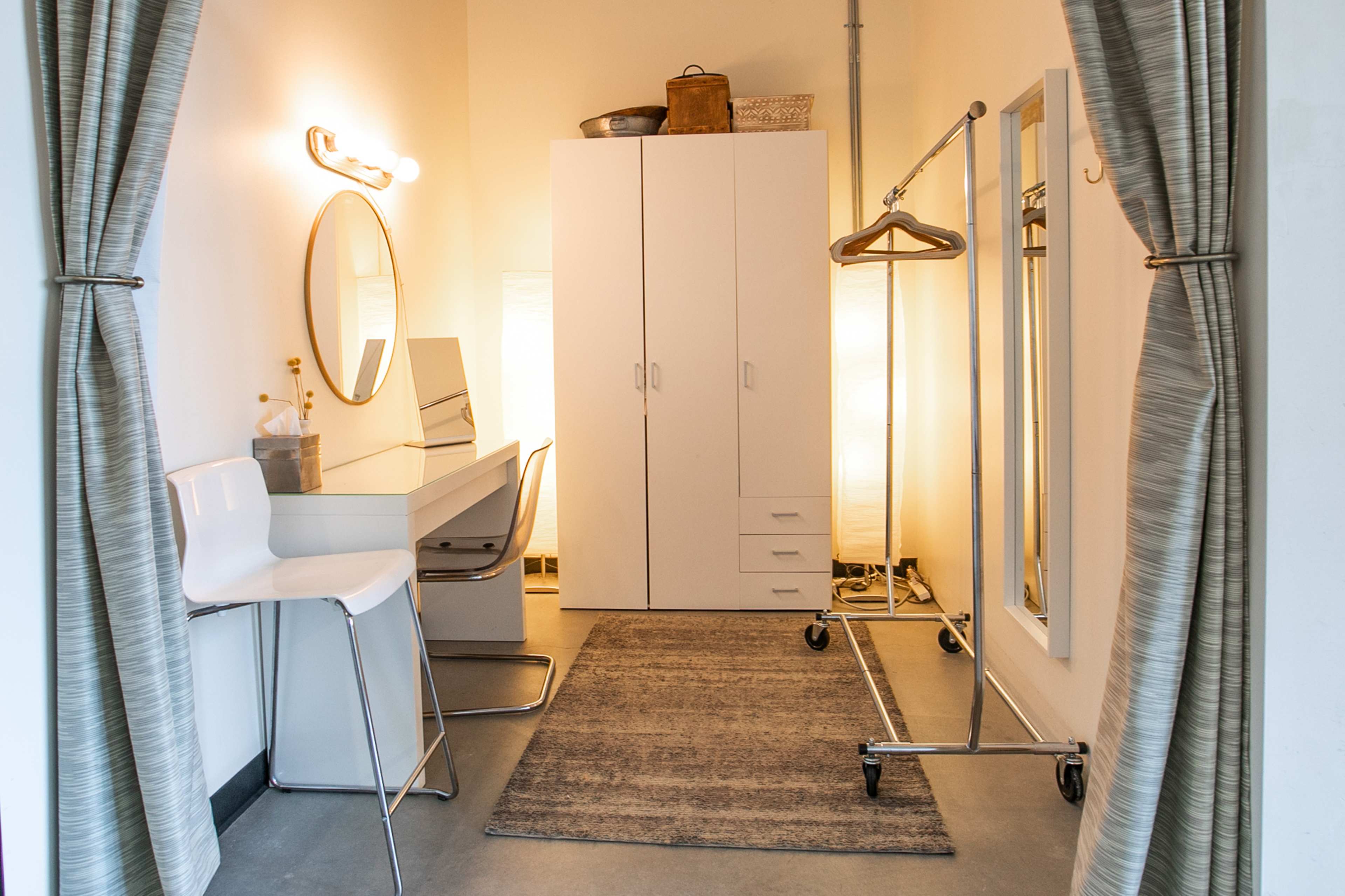 A small dressing room with a mirror, a chair, a clothing rack, and a storage cabinet against the wall, illuminated by soft lighting.