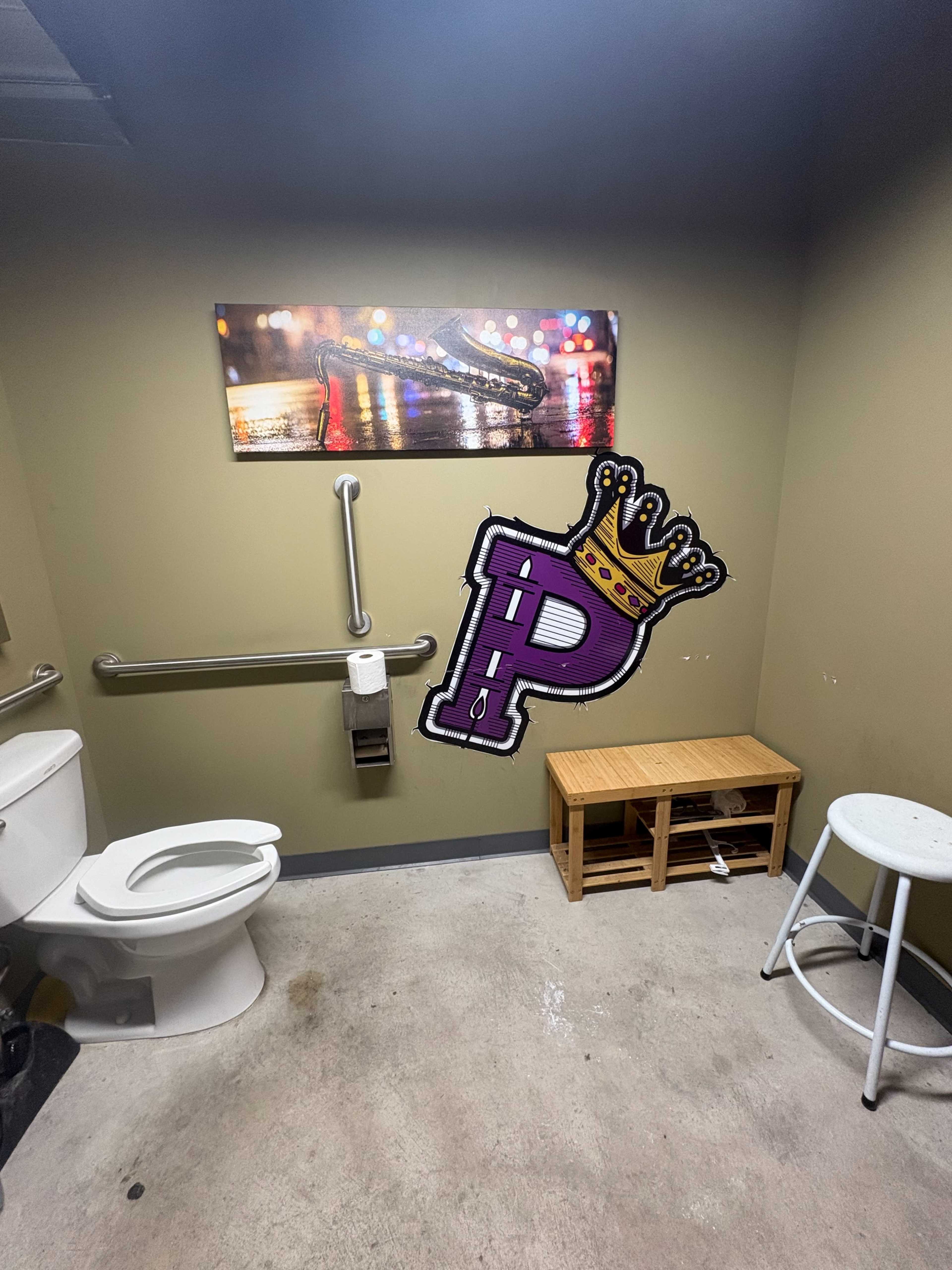 The image shows a small restroom with a toilet, a wooden bench, and a wall featuring an artistic representation of a crown and the letter "P."