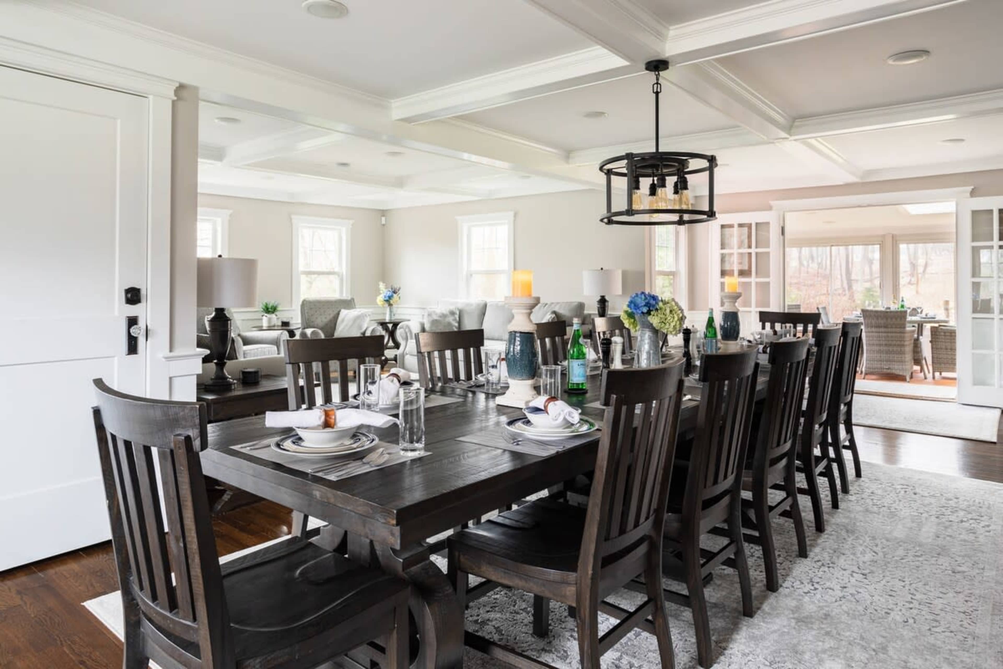 A long wooden dining table is set for a meal, surrounded by dark chairs, with a view into a bright living area through open doors.