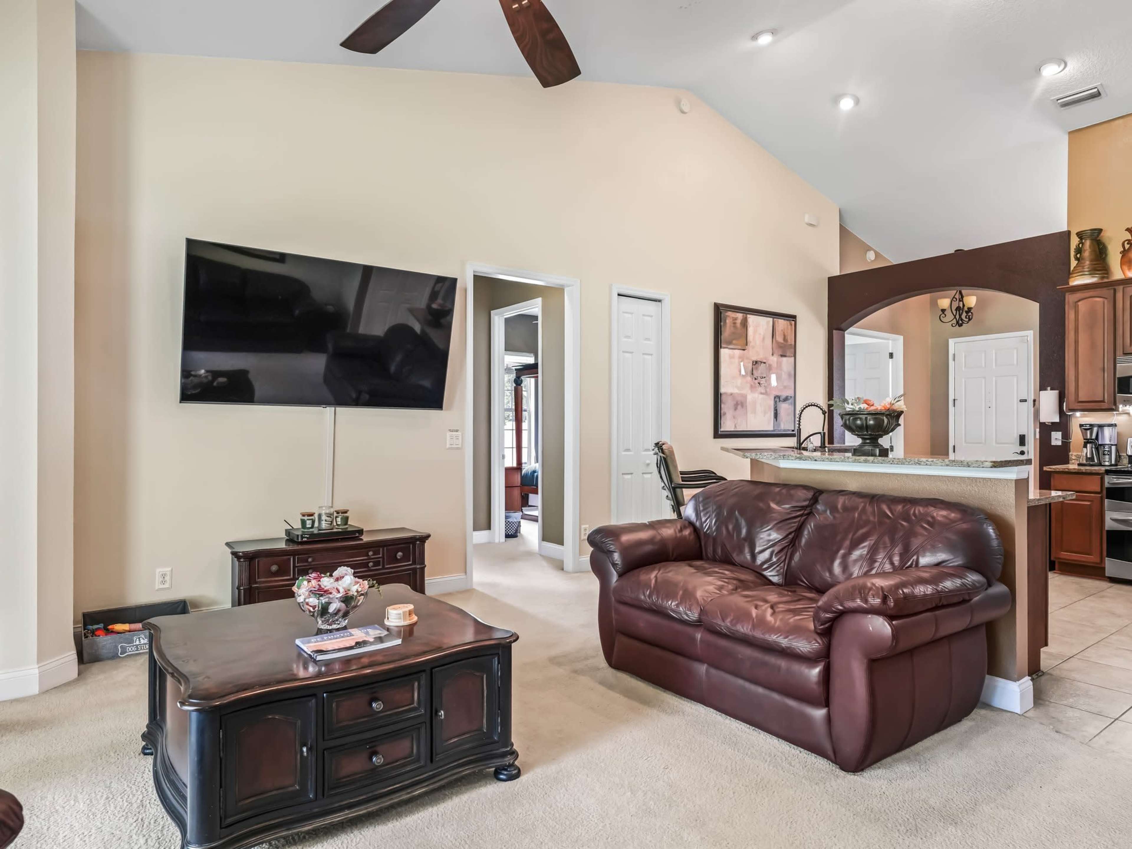 The living area features a leather sofa, a coffee table, and a wall-mounted television, adjacent to an open kitchen space.