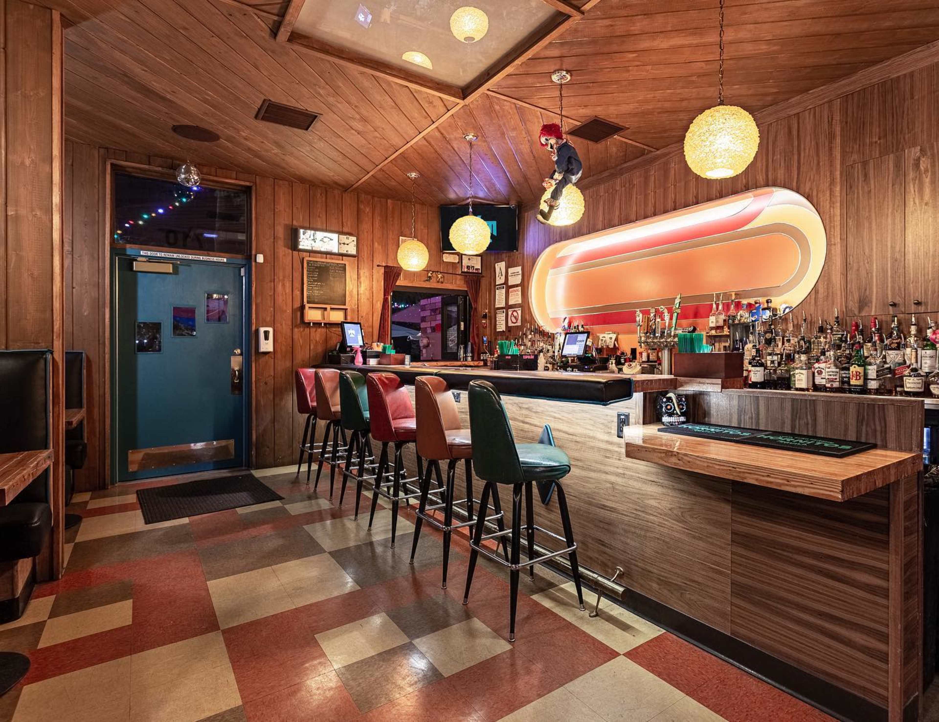 The image shows a bar area with a wooden interior, colorful stools, and a well-stocked bar illuminated by hanging pendant lights.