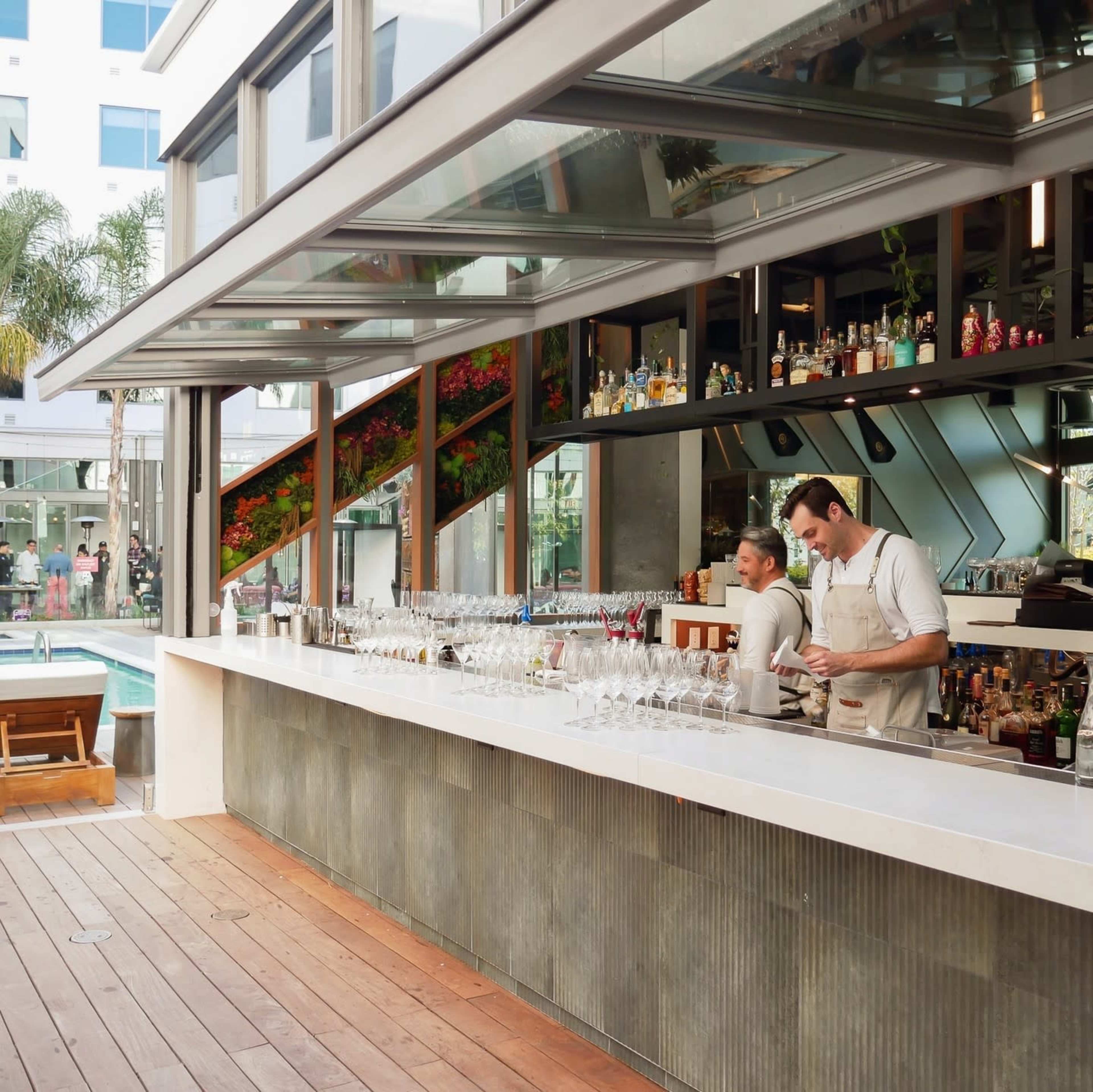 Two bartenders are preparing drinks at a modern bar with a large outdoor seating area, surrounded by plants and a swimming pool.
