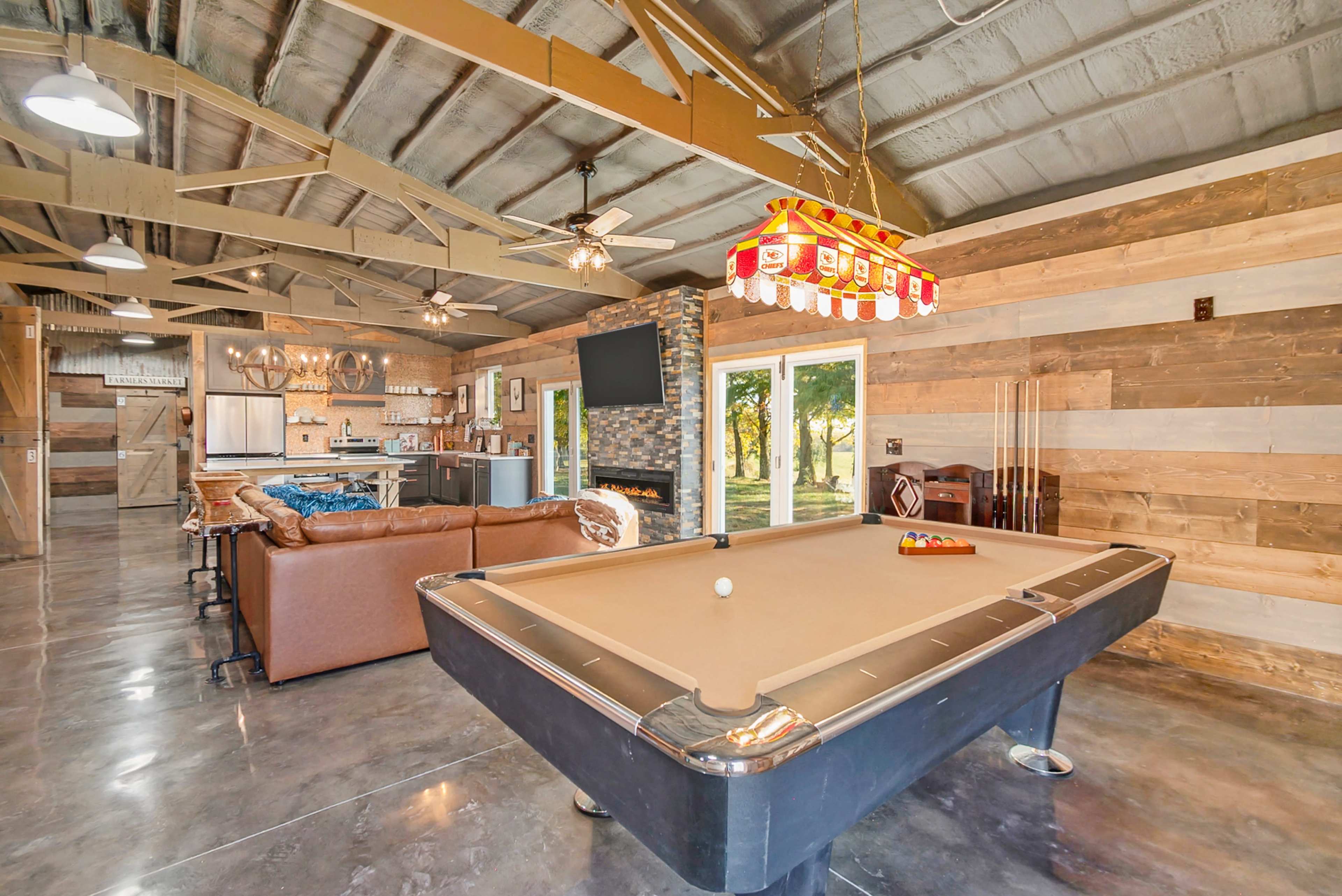 The image shows a spacious game room featuring a pool table, a leather sofa, a flat-screen TV, and large windows that provide a view of the outdoors.