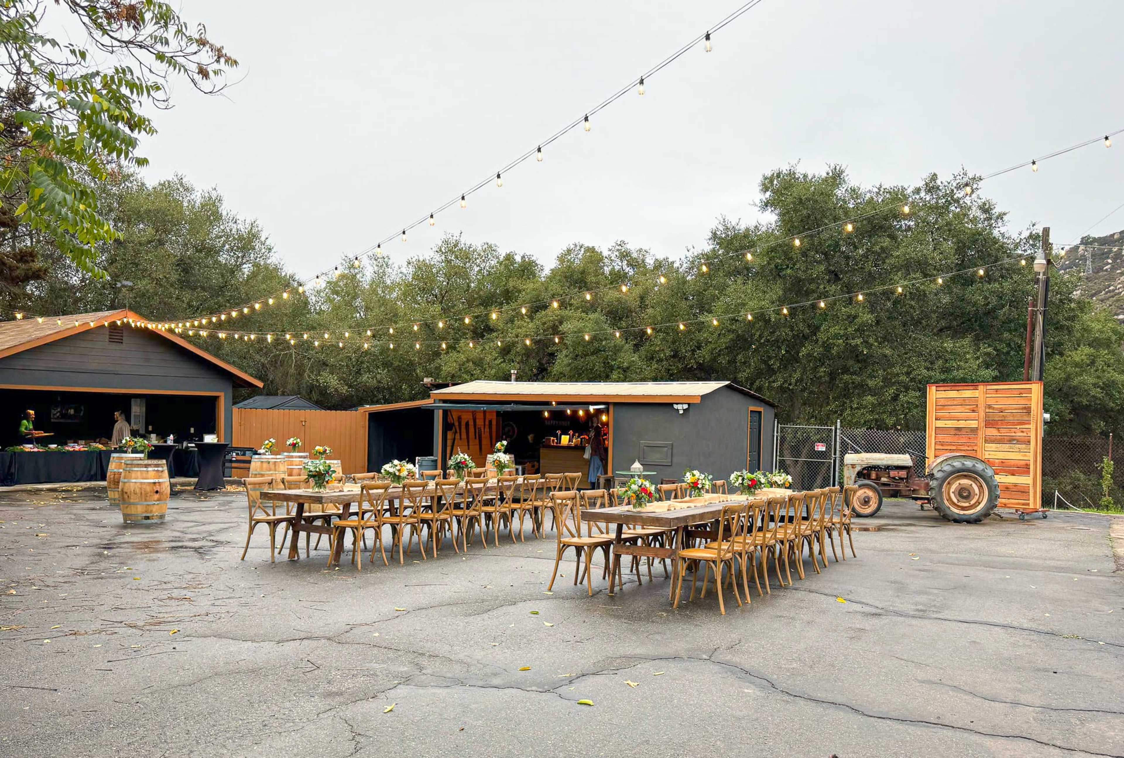 An outdoor event space features long wooden tables adorned with centerpieces, string lights overhead, and a rustic setting with trees and buildings in the background.
