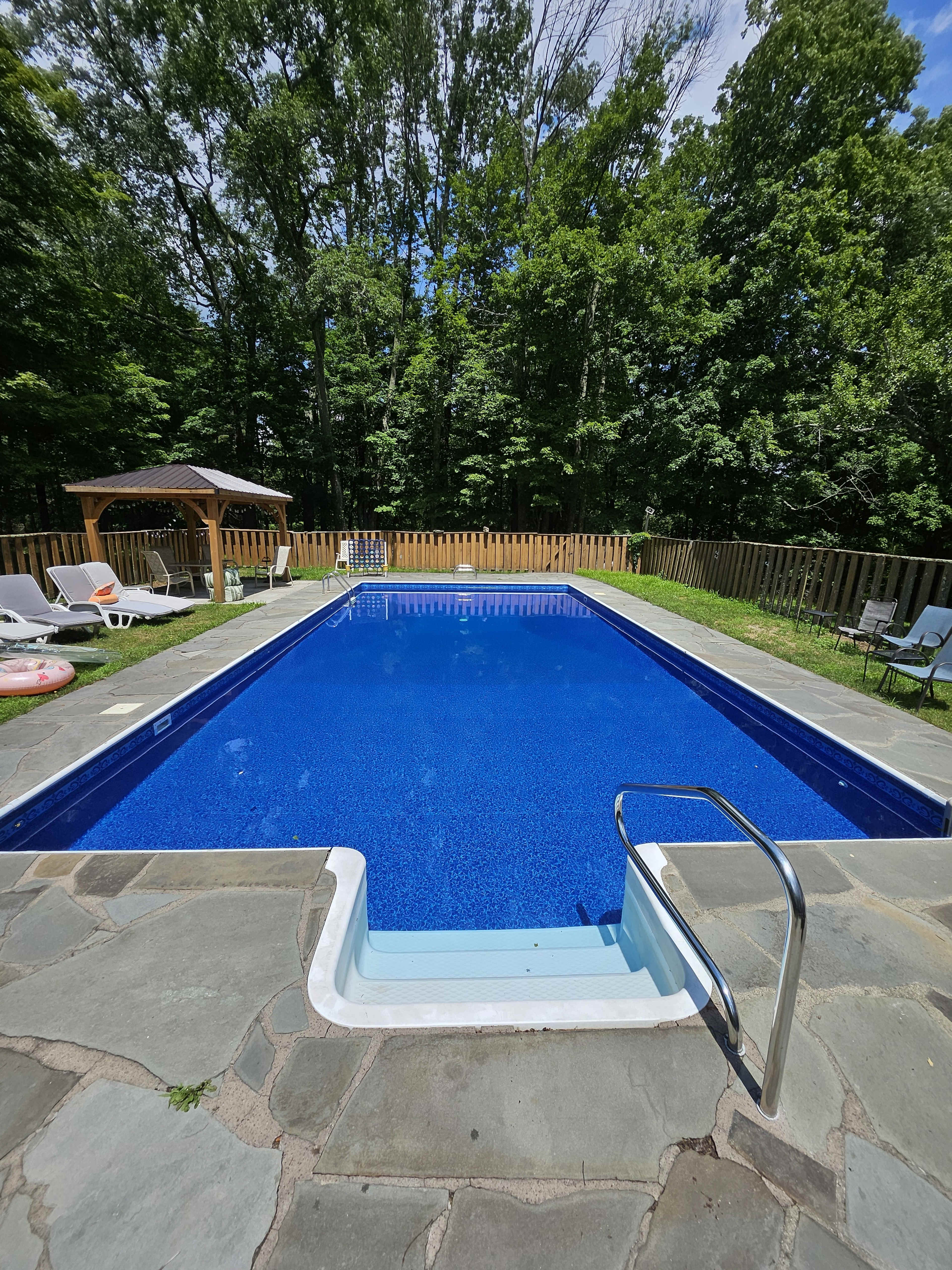 The image shows a rectangular swimming pool surrounded by lush green trees and a wooden fence, with lounge chairs and a gazebo nearby.