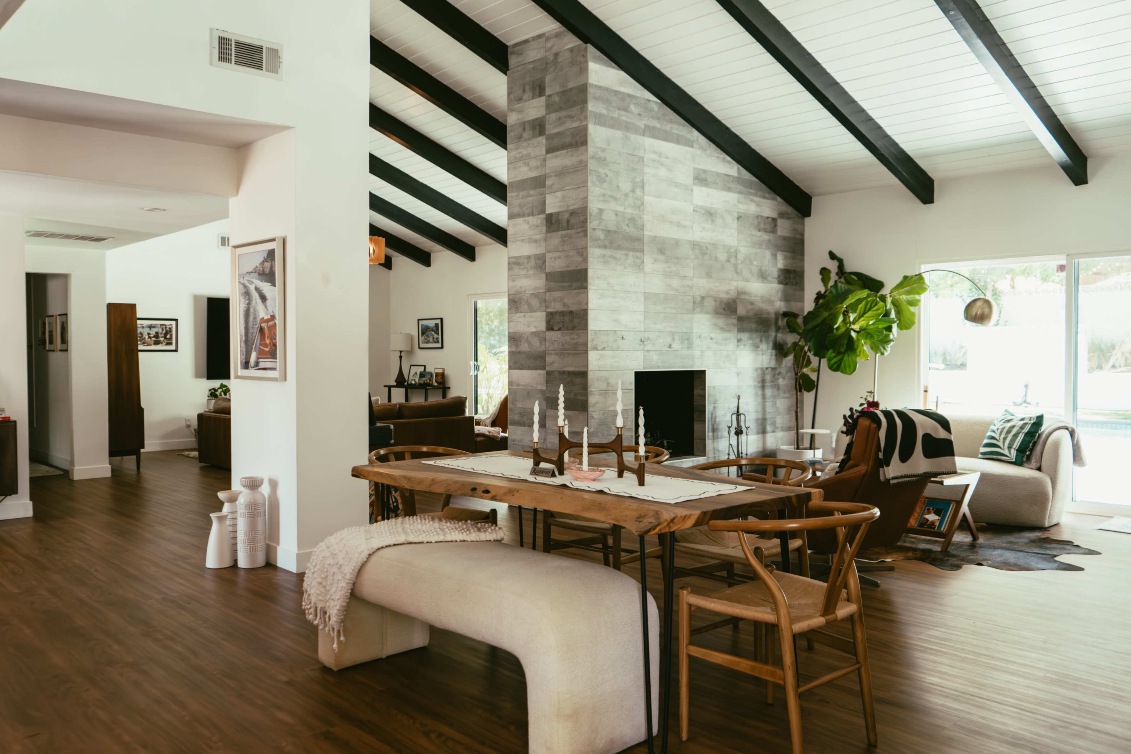 The image shows a modern open-concept living area featuring a wooden dining table, a stone fireplace, and large windows that open to an outdoor space.