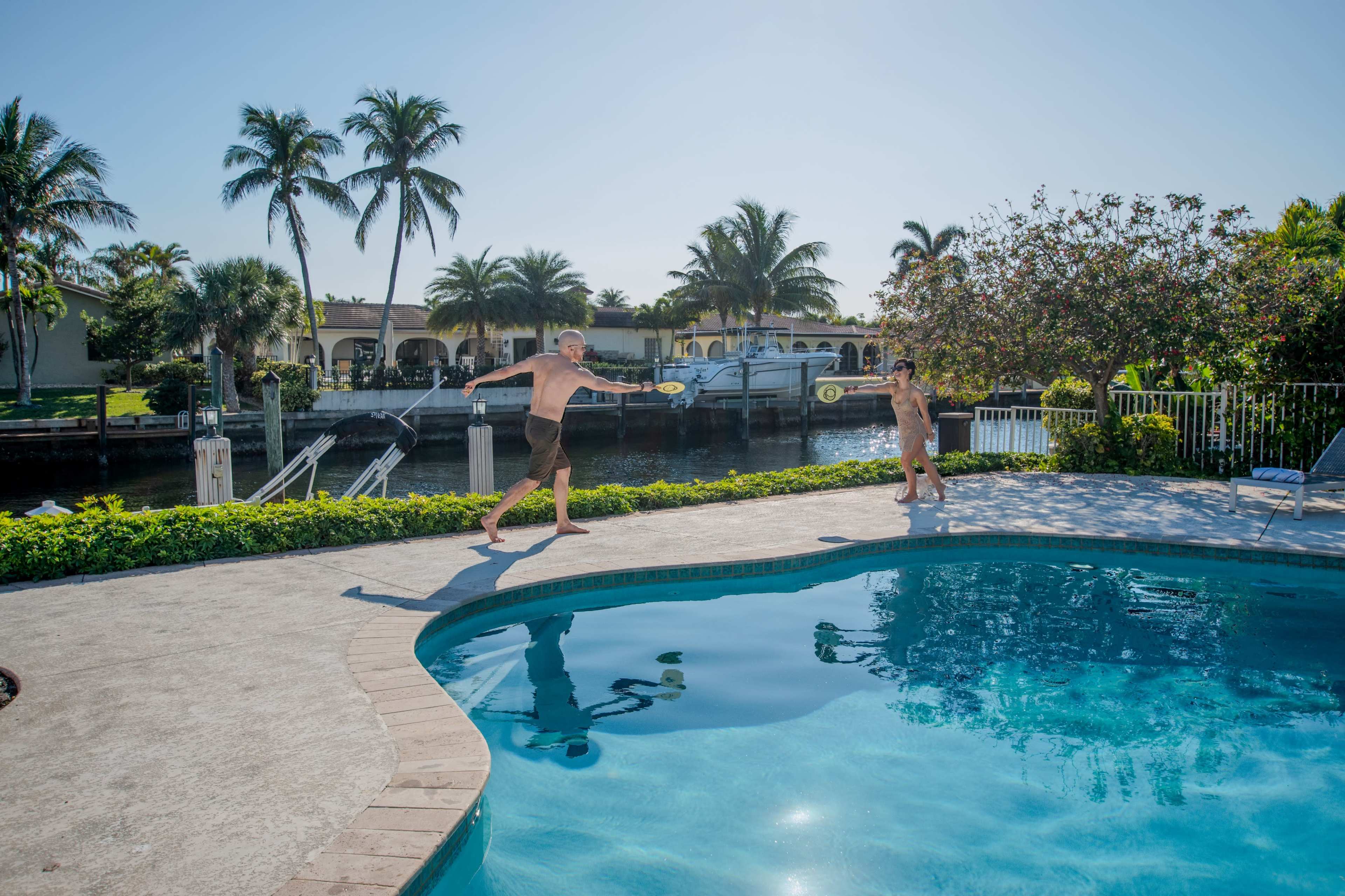 Two children are playing near a swimming pool in a sunny backyard surrounded by palm trees and a waterfront.