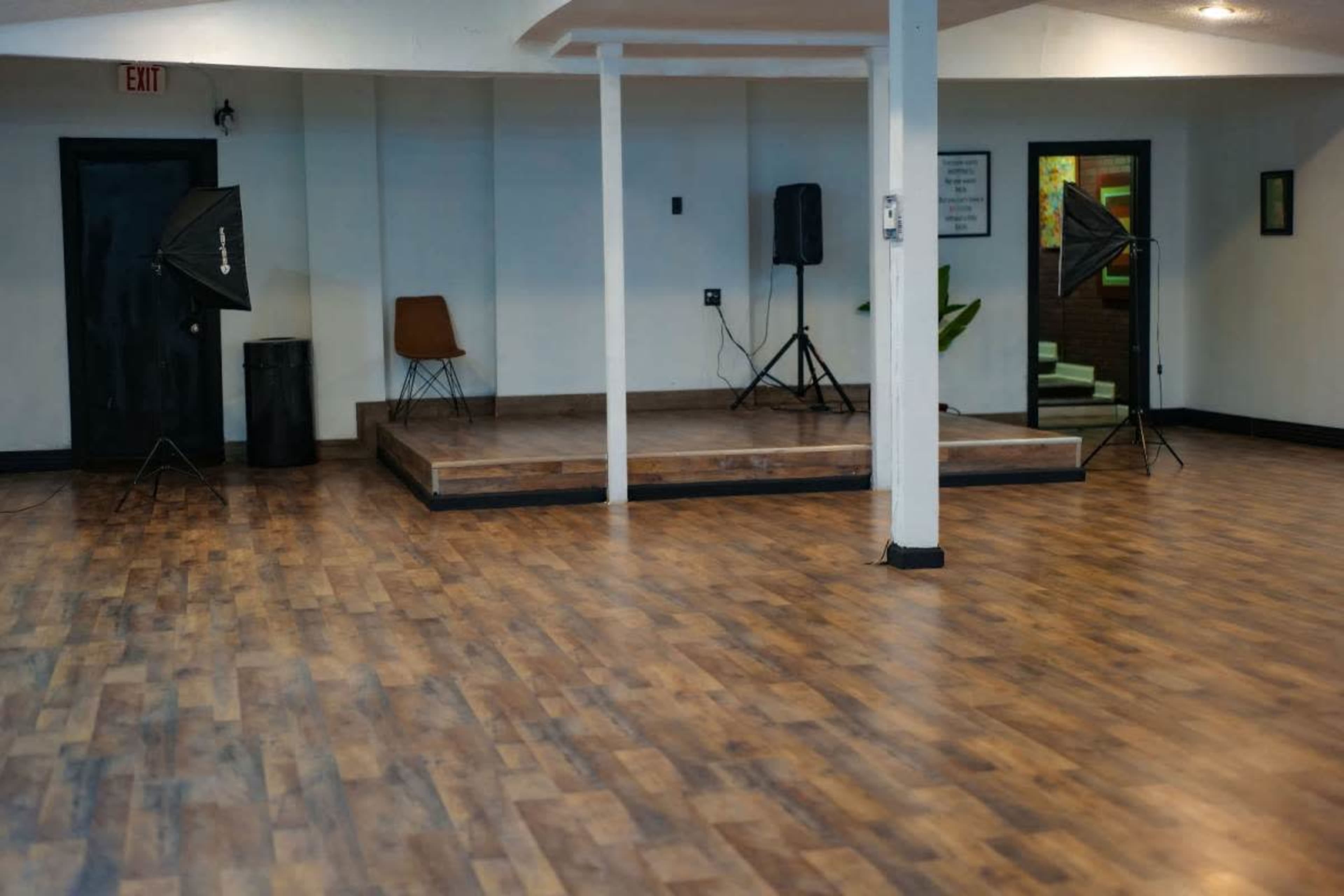 The image shows a spacious, empty room featuring a wooden floor, a small stage with audio equipment, two lighting setups, a chair, and a trash can.
