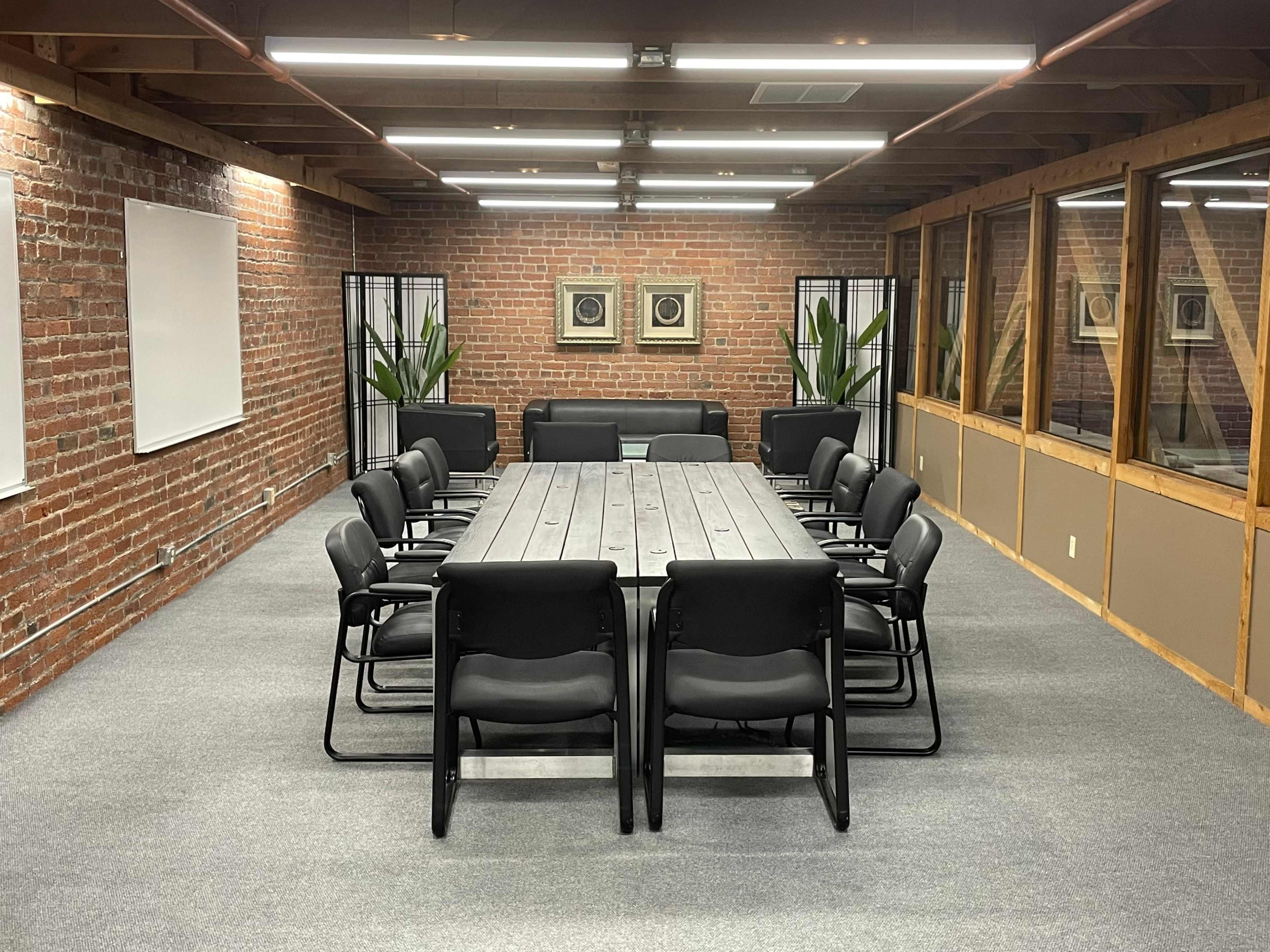 A large conference room features a long wooden table surrounded by black chairs, with exposed brick walls and plants in the corners.
