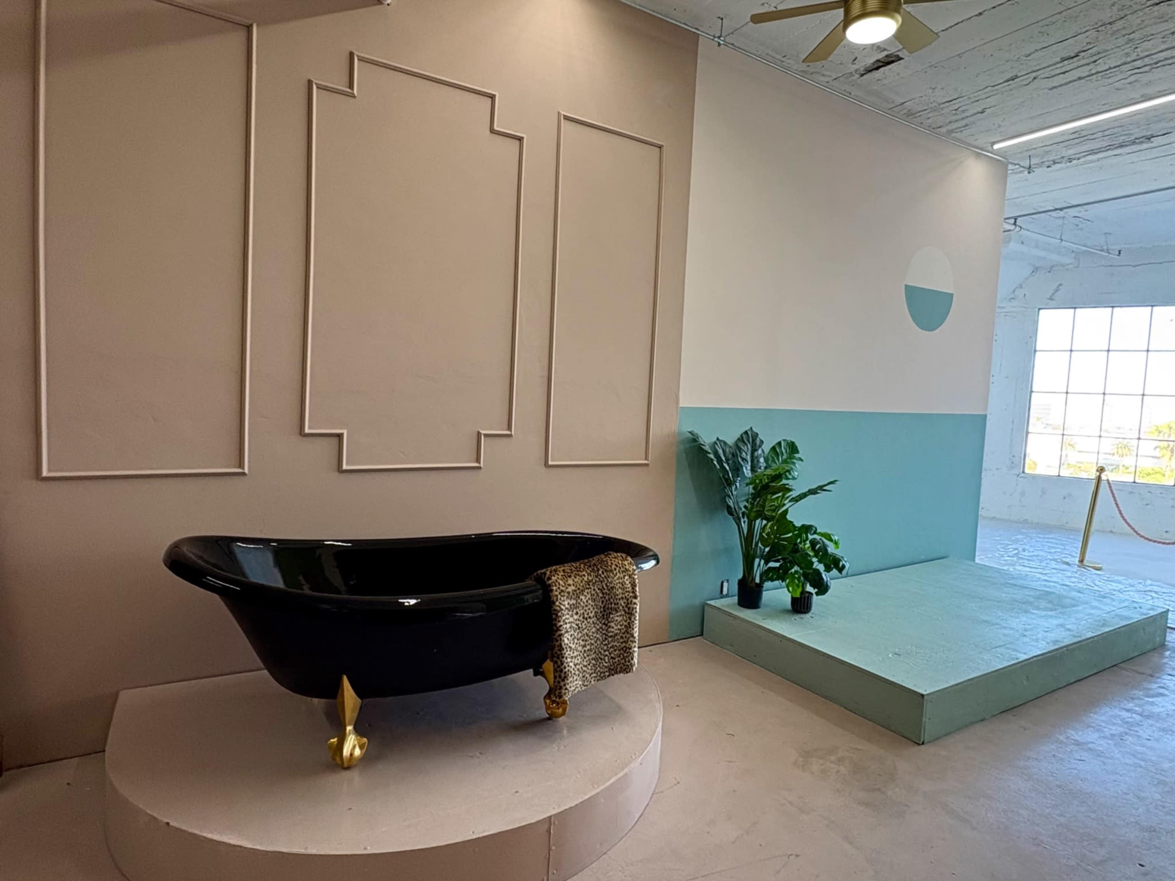 The image depicts a modern bathroom space featuring a black freestanding bathtub on a circular pedestal, set against pastel-colored walls and a corner with greenery.