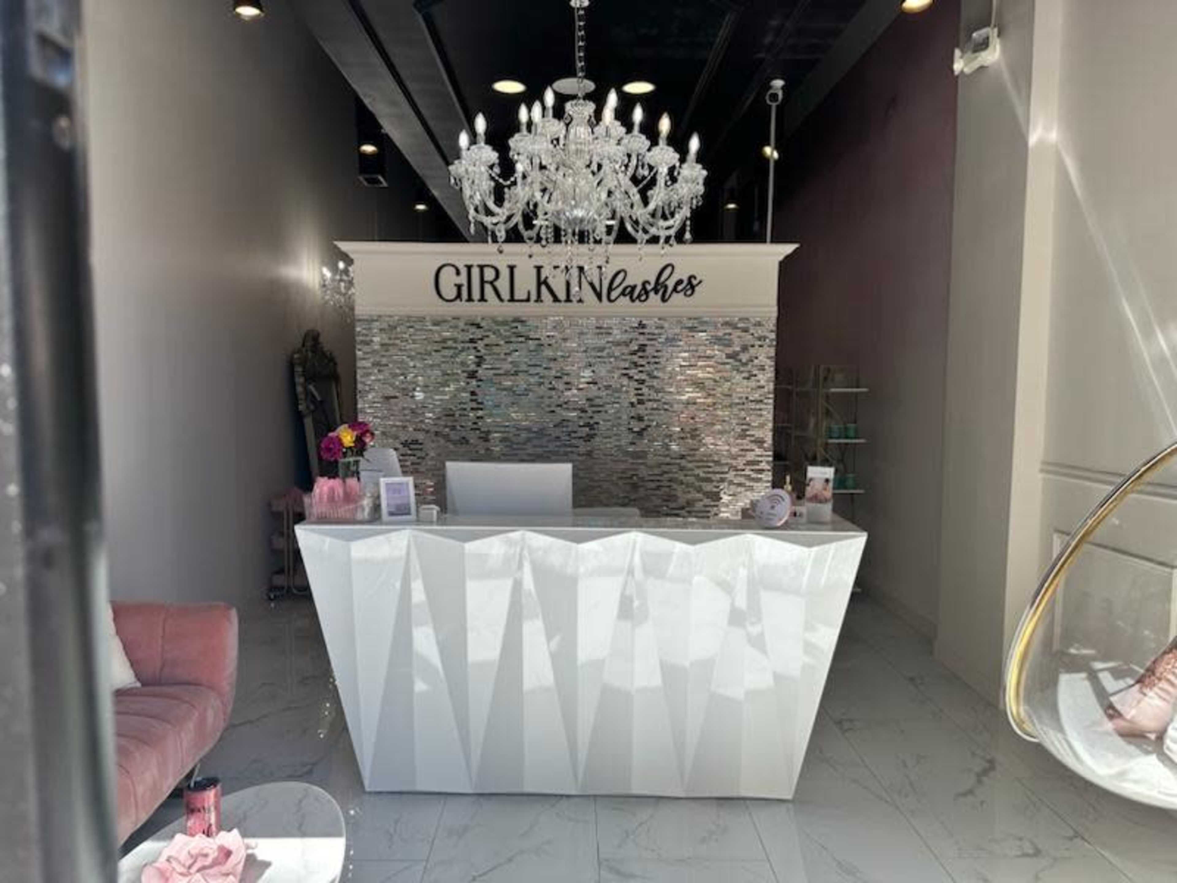 A contemporary lash salon with a reception area featuring a white, geometric front desk and a chandelier overhead.