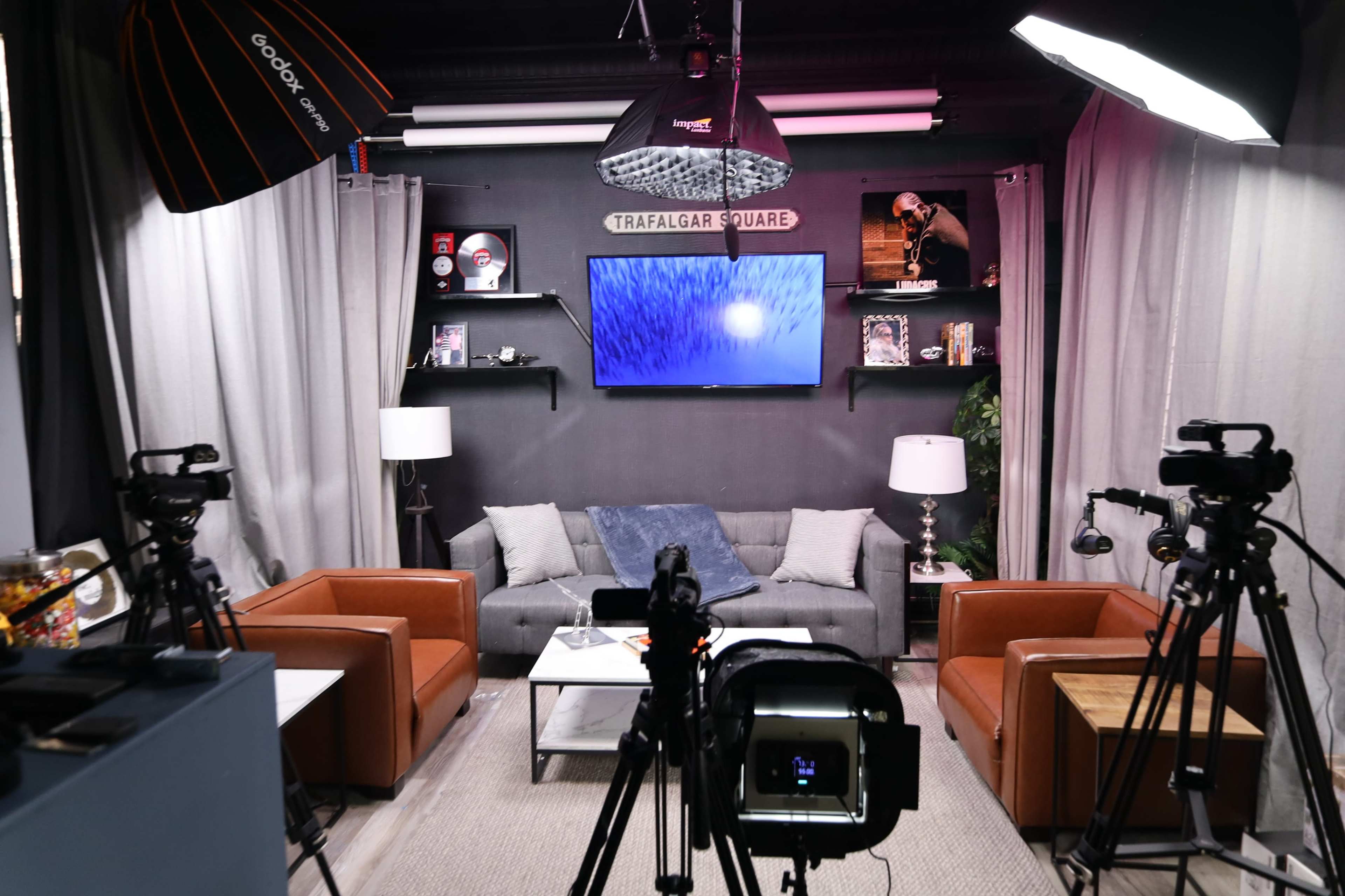 A modern studio setup with two cameras aimed at a seating area featuring a gray couch, two brown chairs, and a screen displaying visuals on the wall.