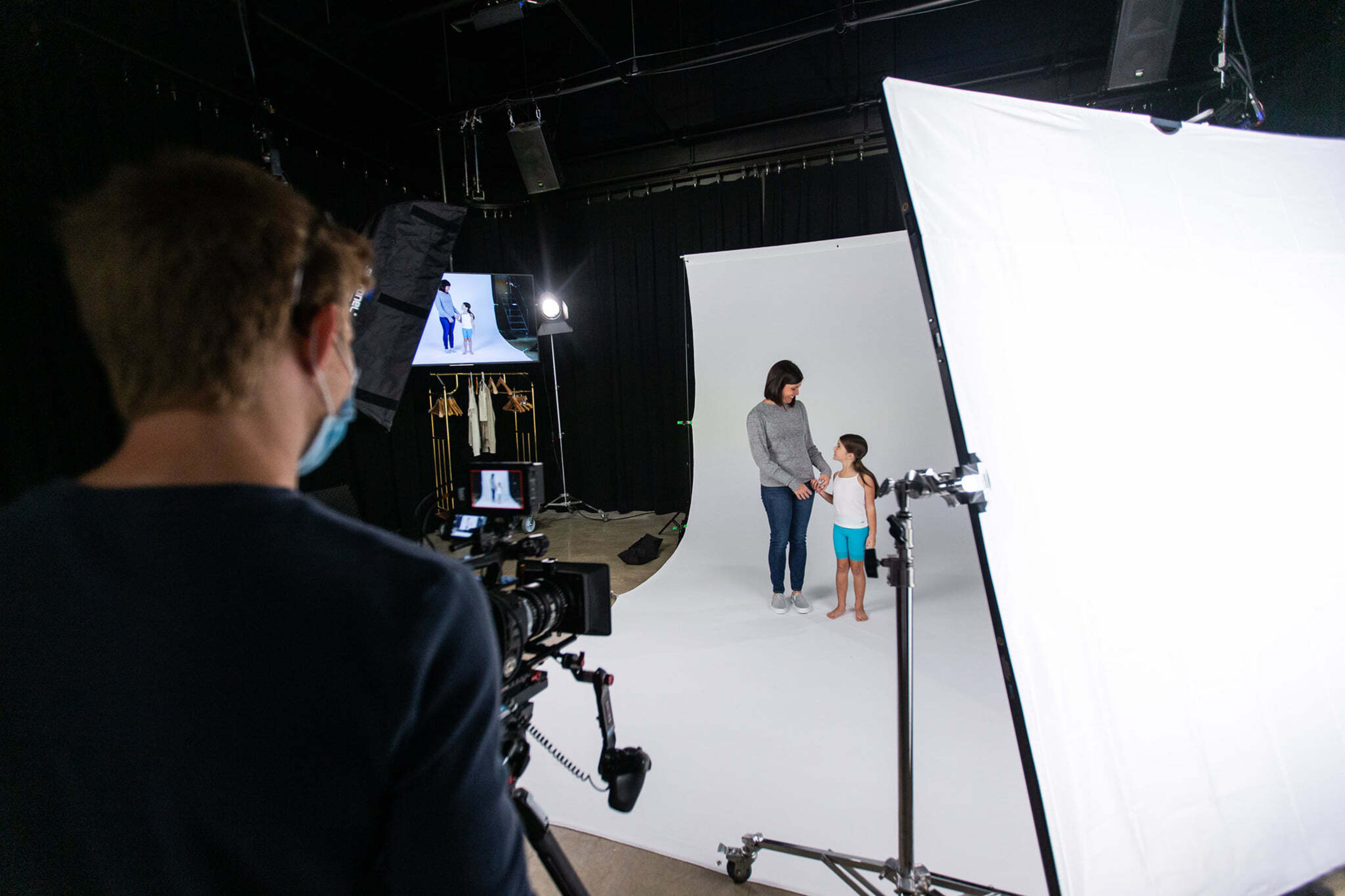 A photographer captures a woman holding hands with a child in a studio setting with a white backdrop and lighting equipment.