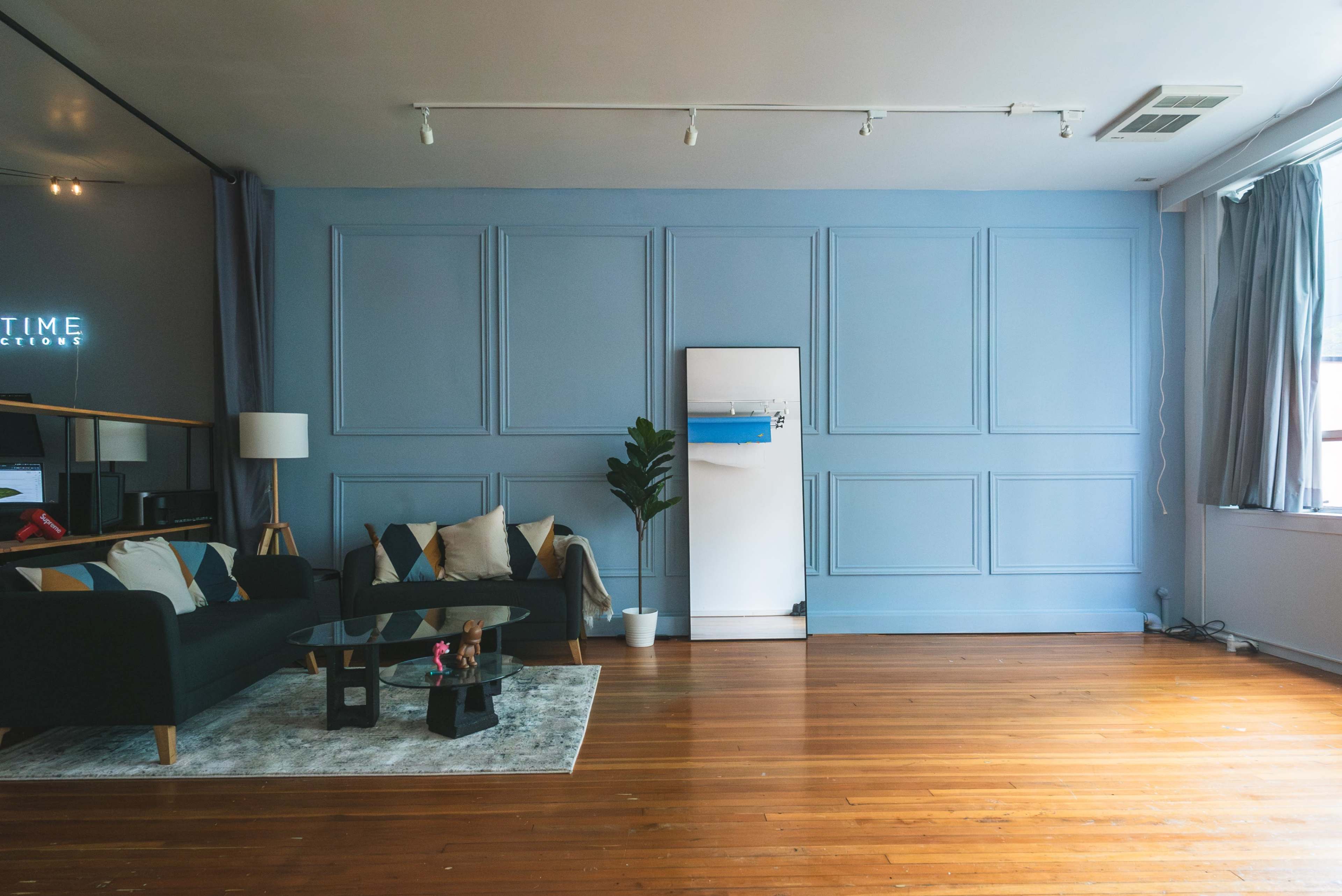 A spacious living room with a green sofa, patterned pillows, a coffee table, a plant in a pot, and a white refrigerator against a blue wall.