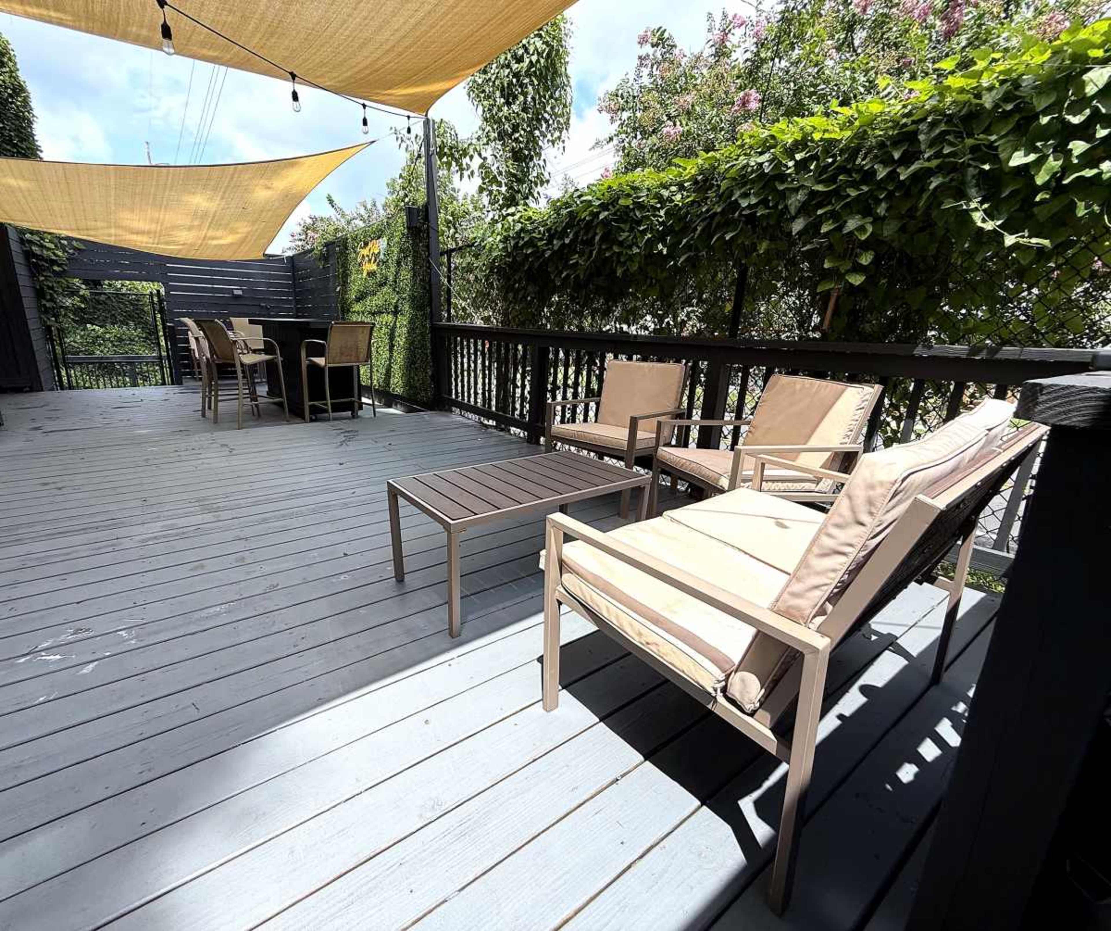 The image depicts a wooden deck featuring two lounge chairs and a table, shaded by fabric canopies and surrounded by lush green foliage.