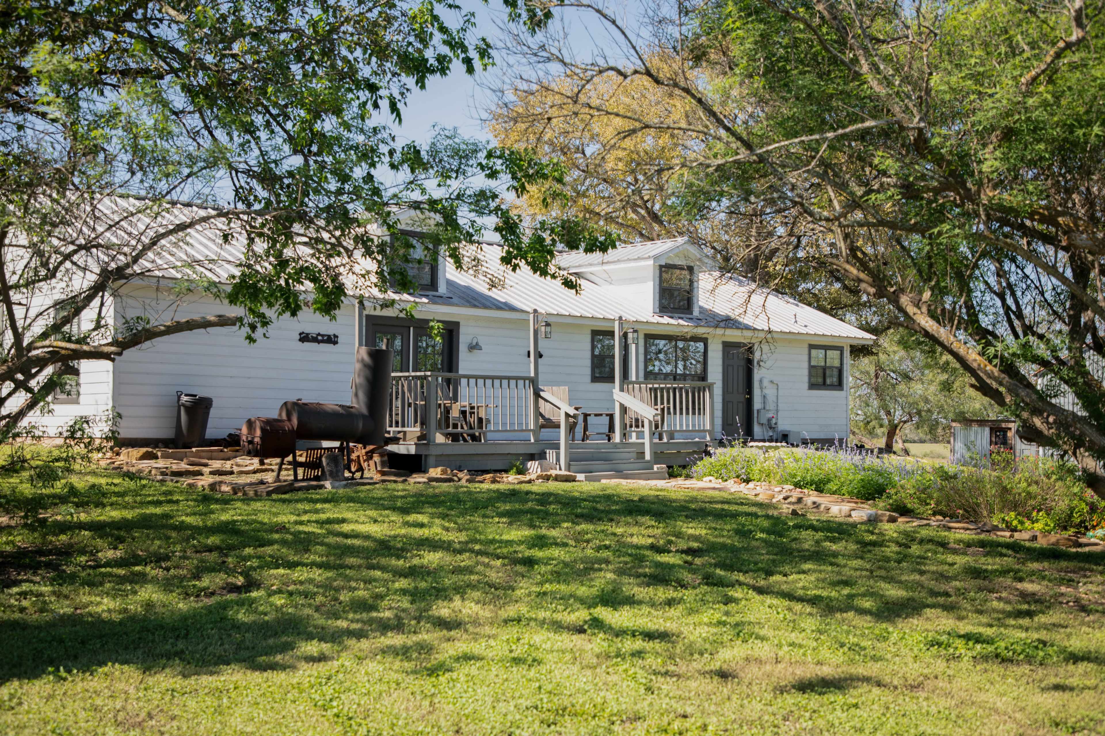 A white, modern farmhouse with a metal roof sits on a grassy lawn, surrounded by trees and flower beds.